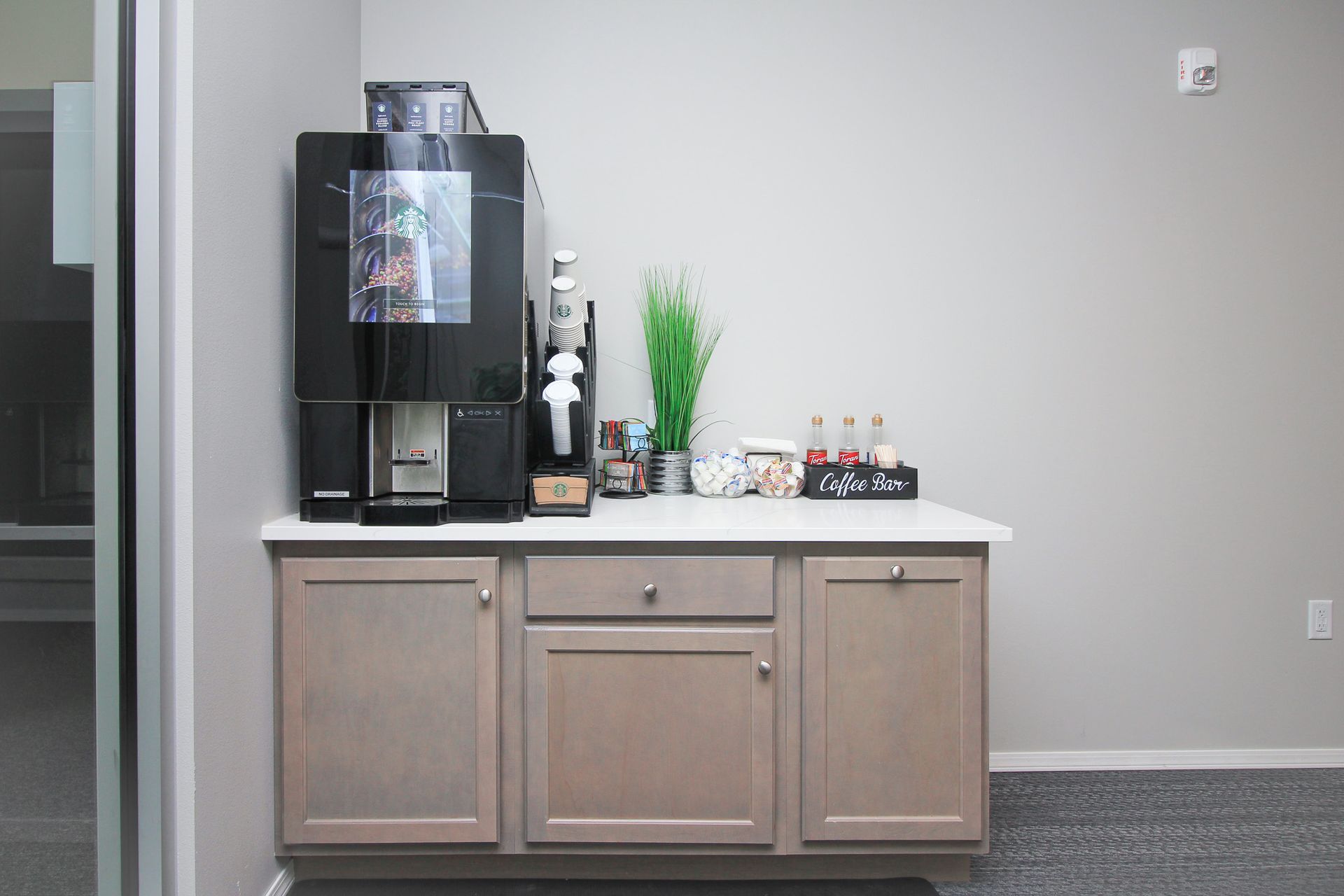 Coffee station with black machine on a light wood cabinet against a white wall.