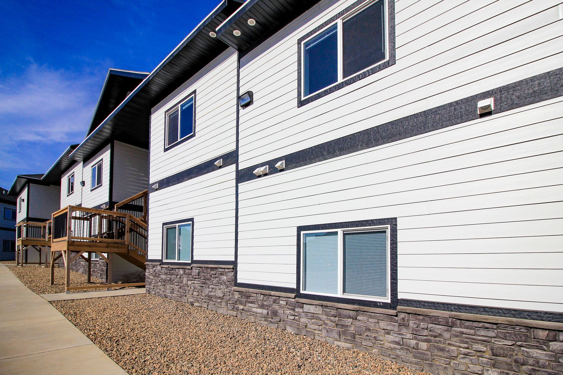 Apartment building exterior with white siding, stone base, and wooden porches under a blue sky