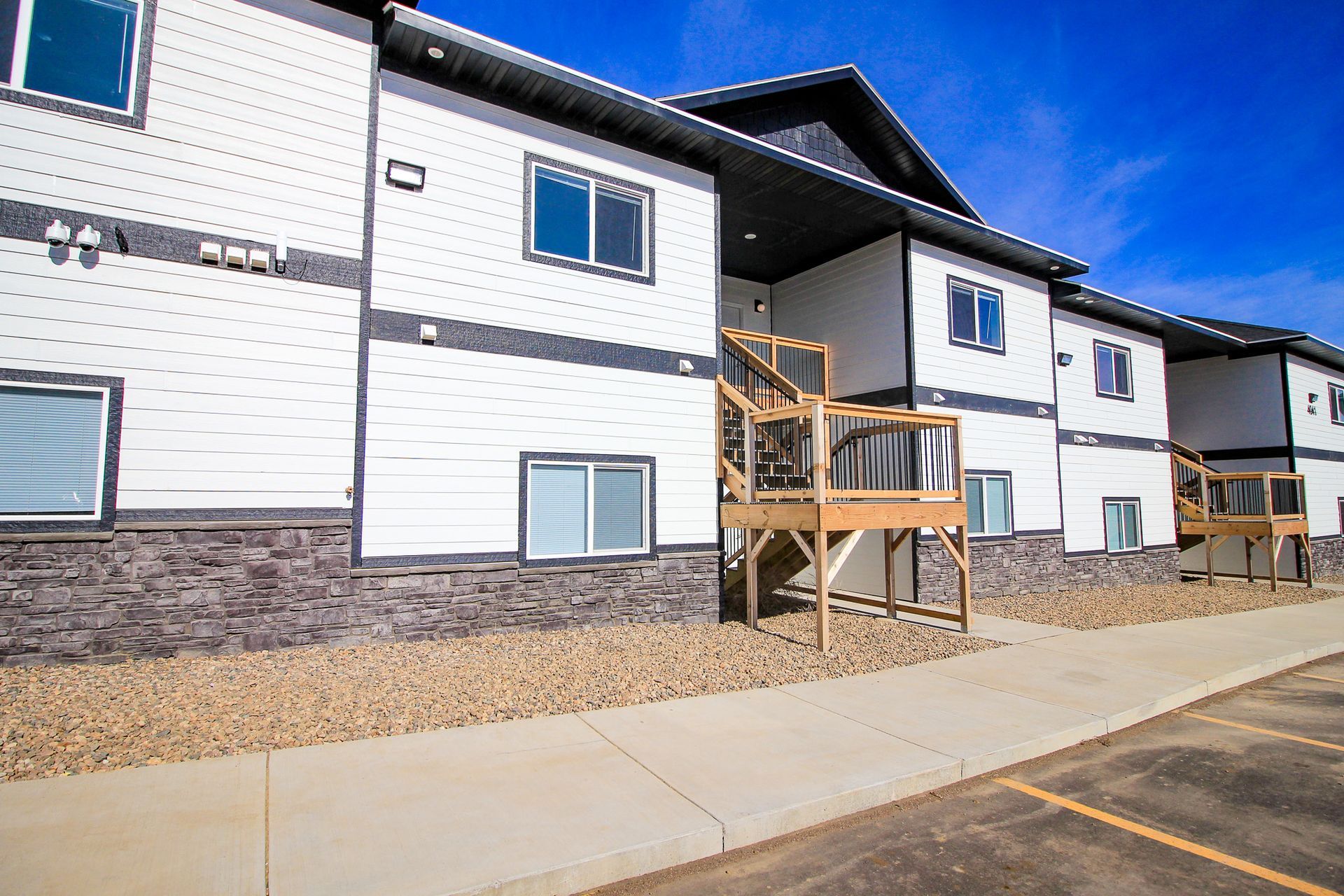 Exterior of a modern apartment building with white siding, stone base, and wooden balconies on a sidewalk