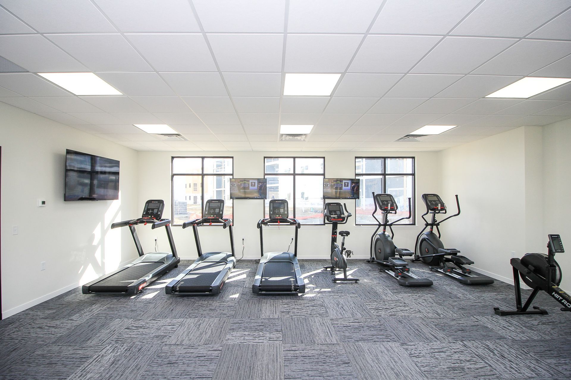 Gym with treadmills, ellipticals, wall TV, and large windows on a gray carpeted floor.