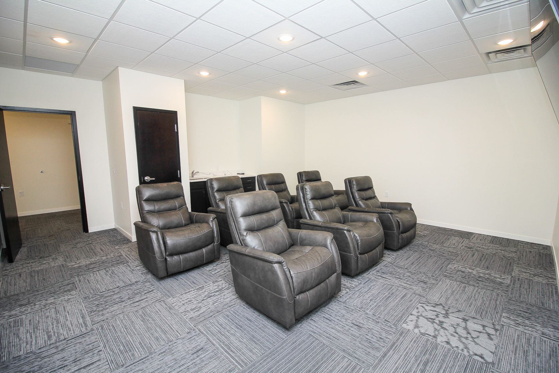 Small lounge with gray recliners, carpeted floor, and white walls under recessed ceiling lights.