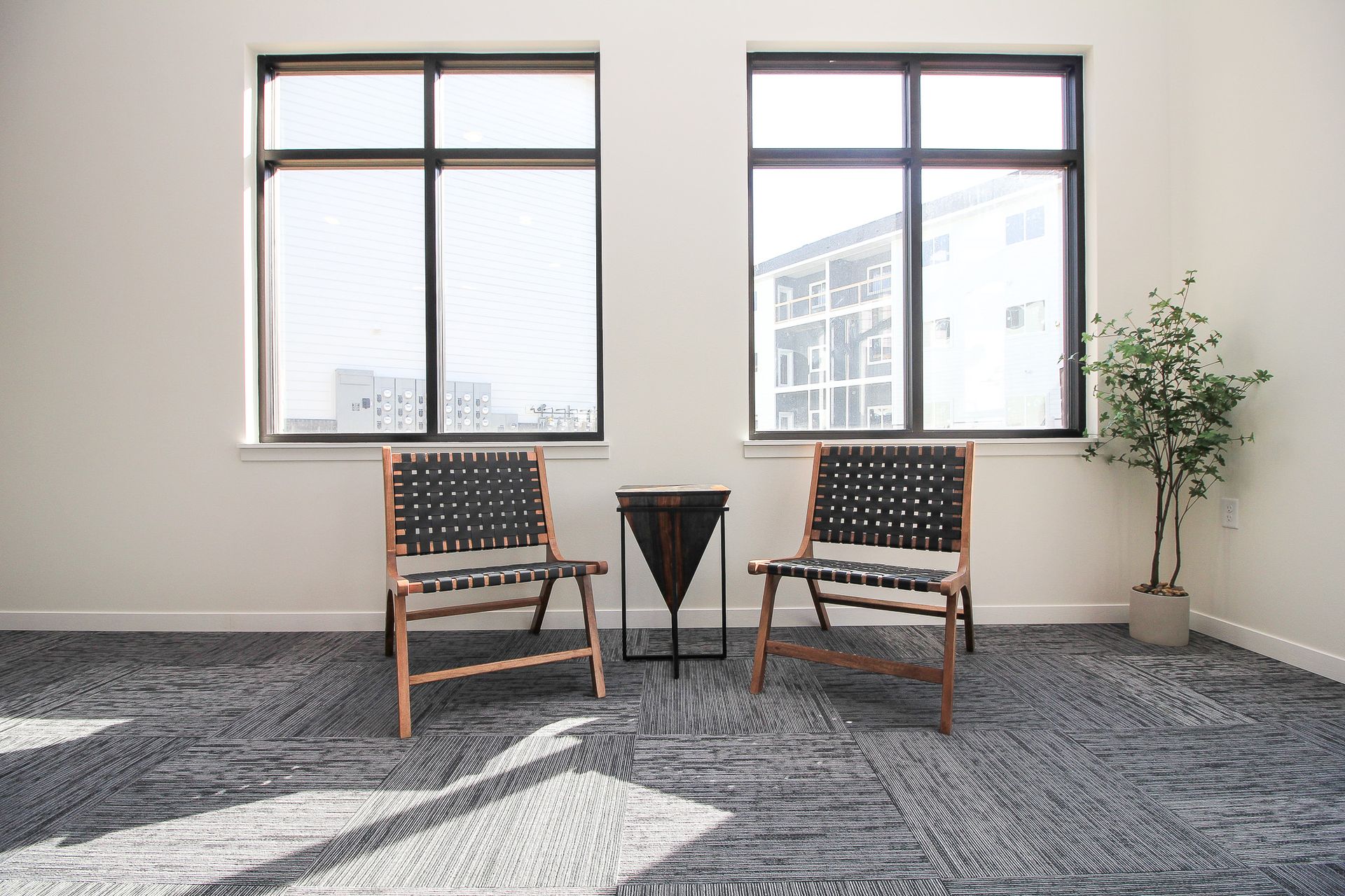 Two wooden chairs beside a small black table in a bright room with windows and a potted plant.