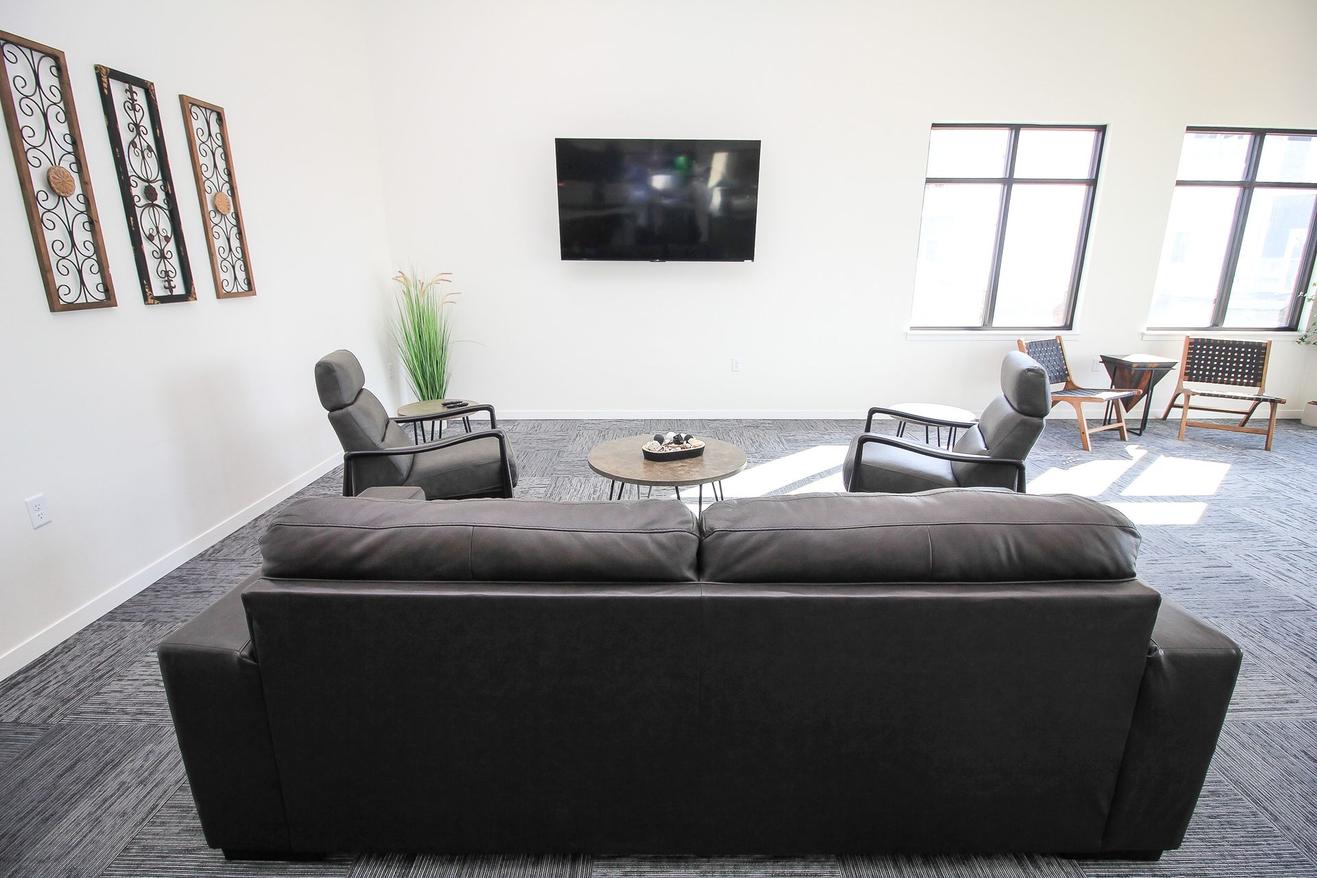 Modern living room with black sofa, wall-mounted TV, gray chairs, and bright windows