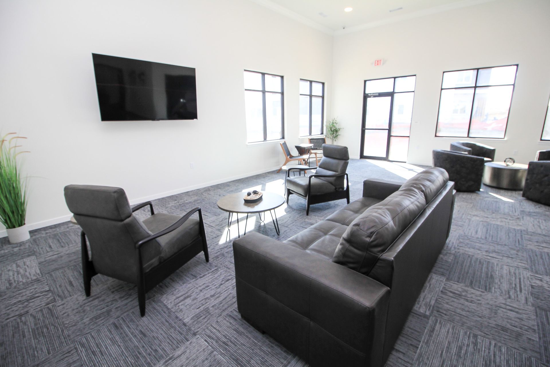 Modern lounge with dark sofas, armchair, wall-mounted TV, and large windows on a gray carpeted floor