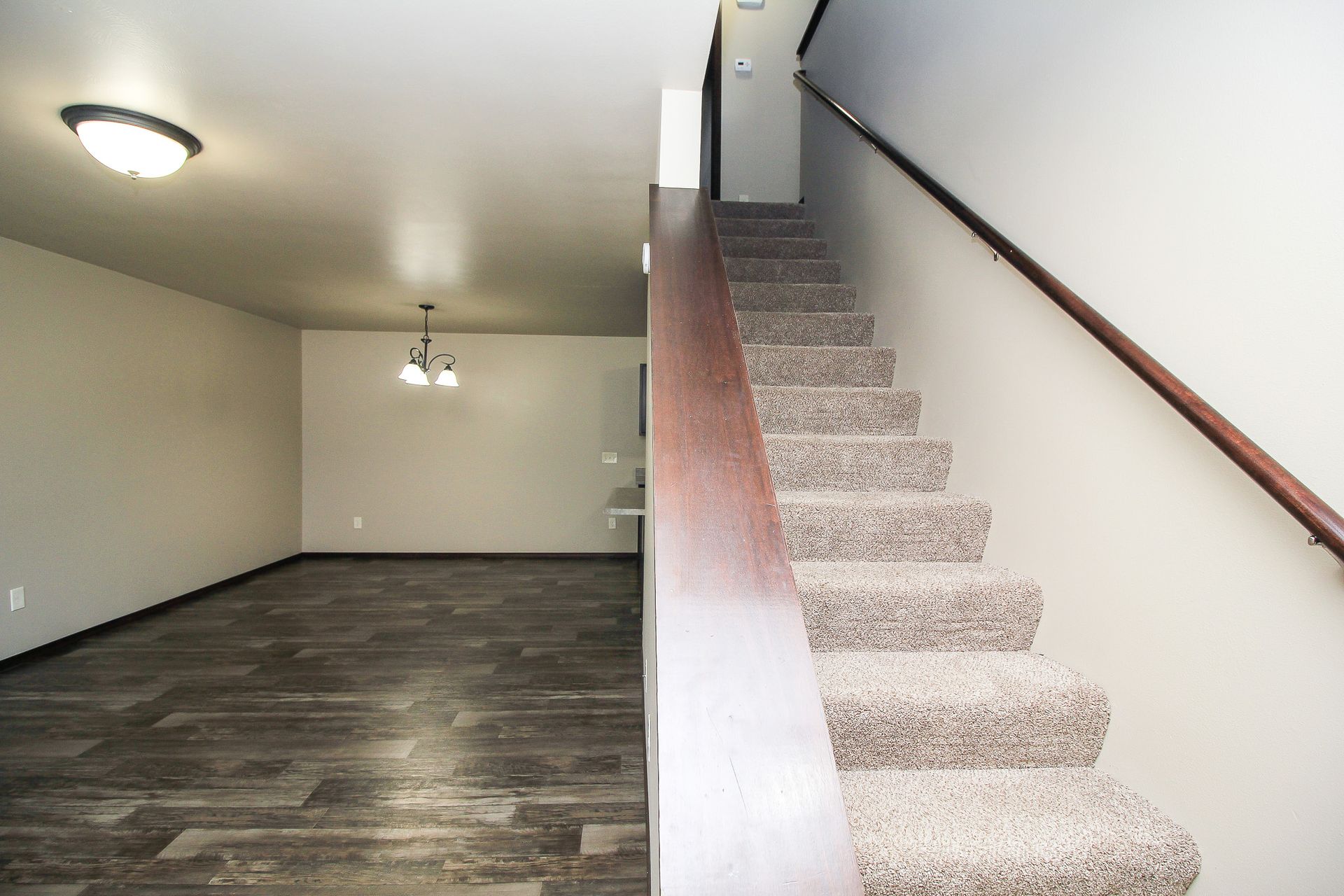 Empty living room beside carpeted staircase with beige walls and dark wood floor.