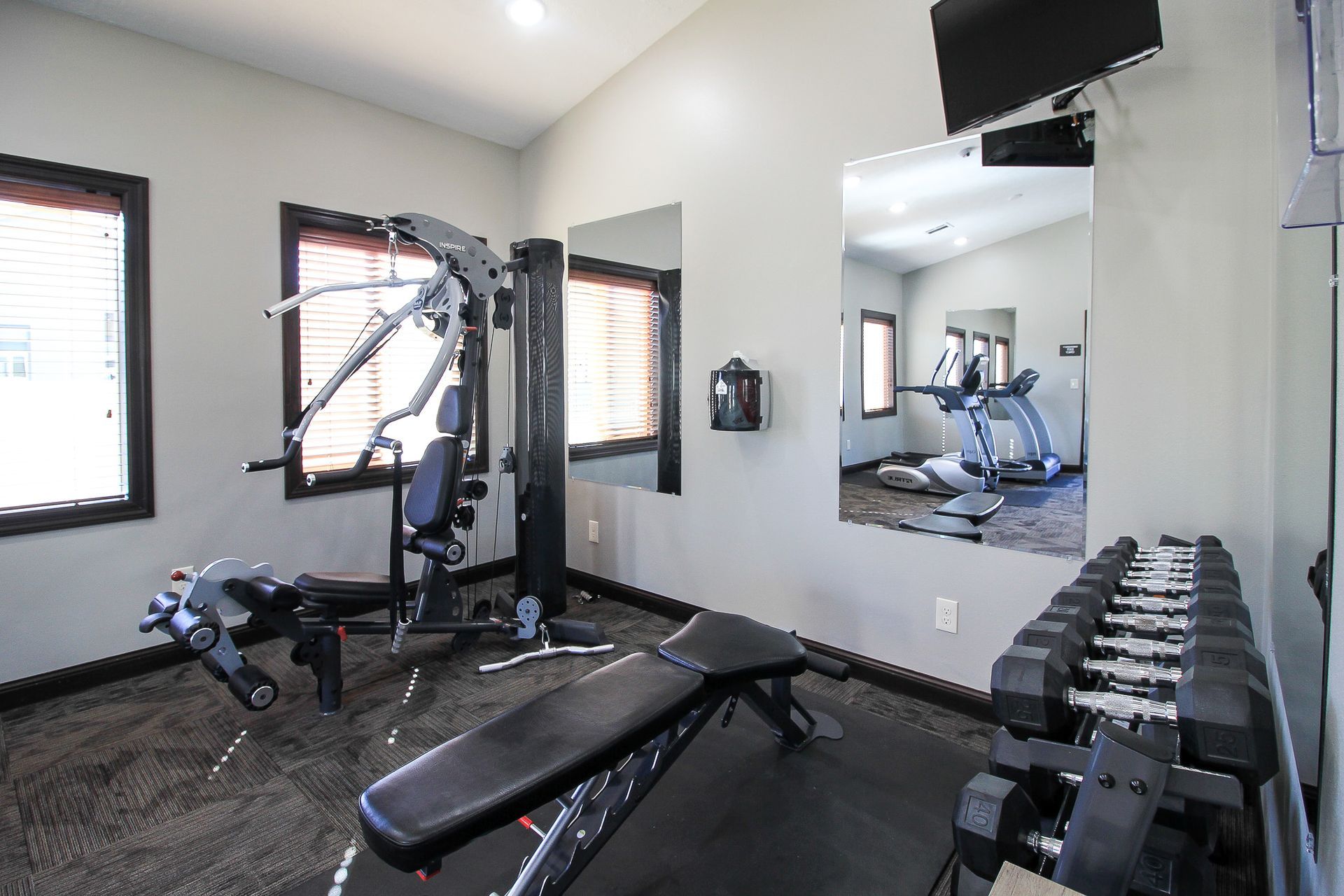 Bright gym room with weight bench, dumbbells, cable machine, mirrors, and windows