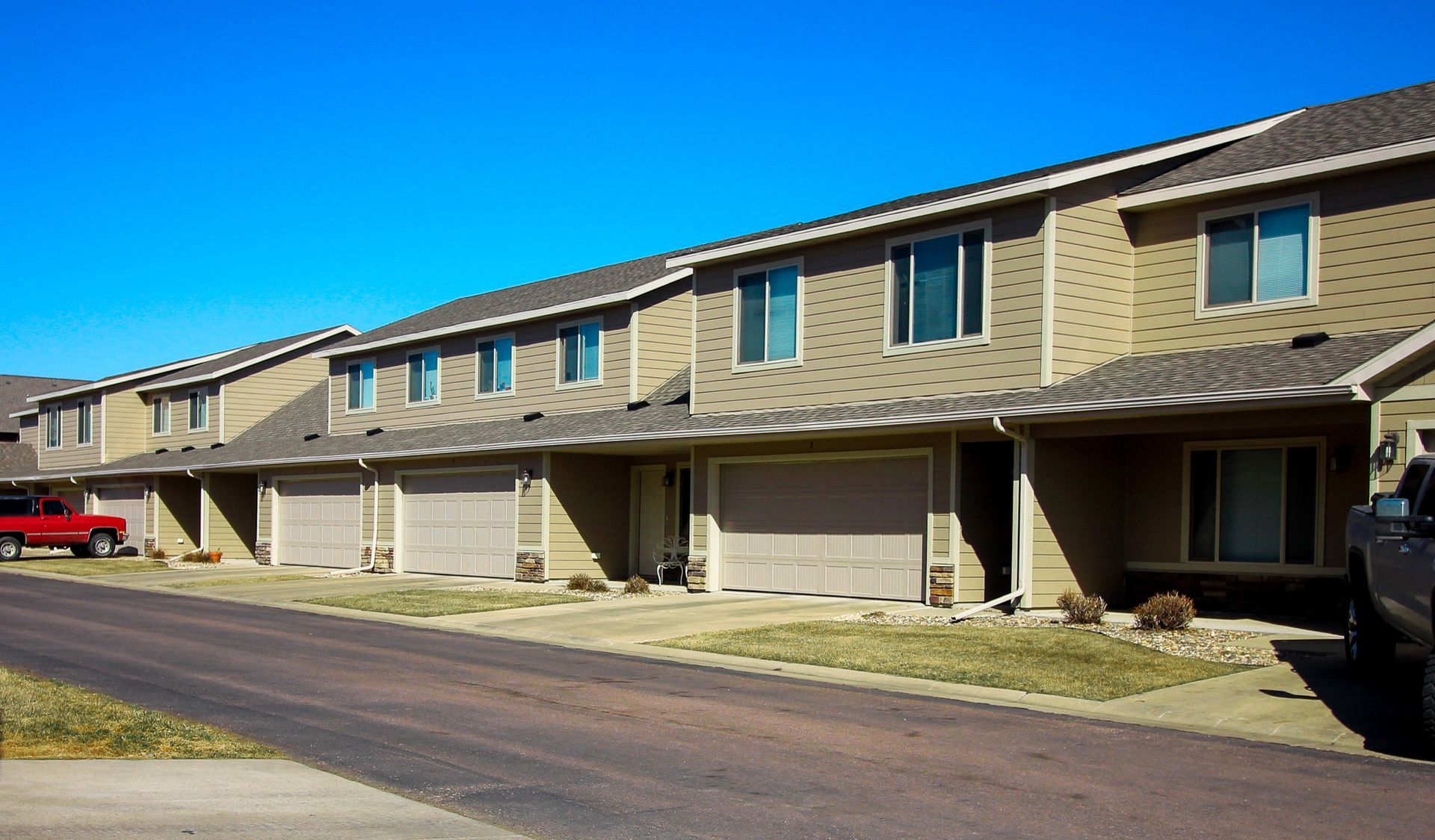Row of tan townhouses with garages along a quiet residential street under a clear blue sky