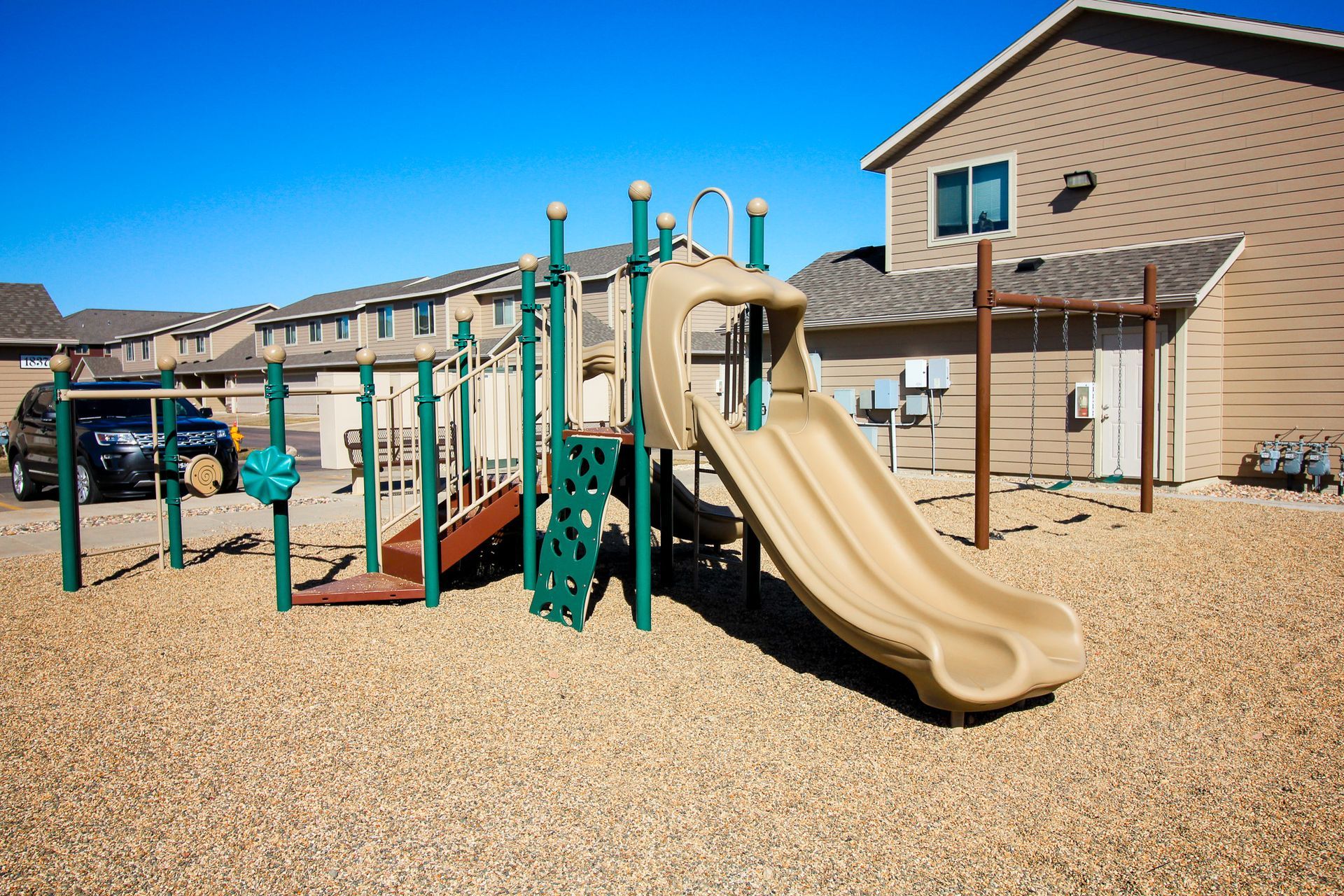 Playground with tan slides and green climbing bars on a gravel-covered schoolyard under a blue sky