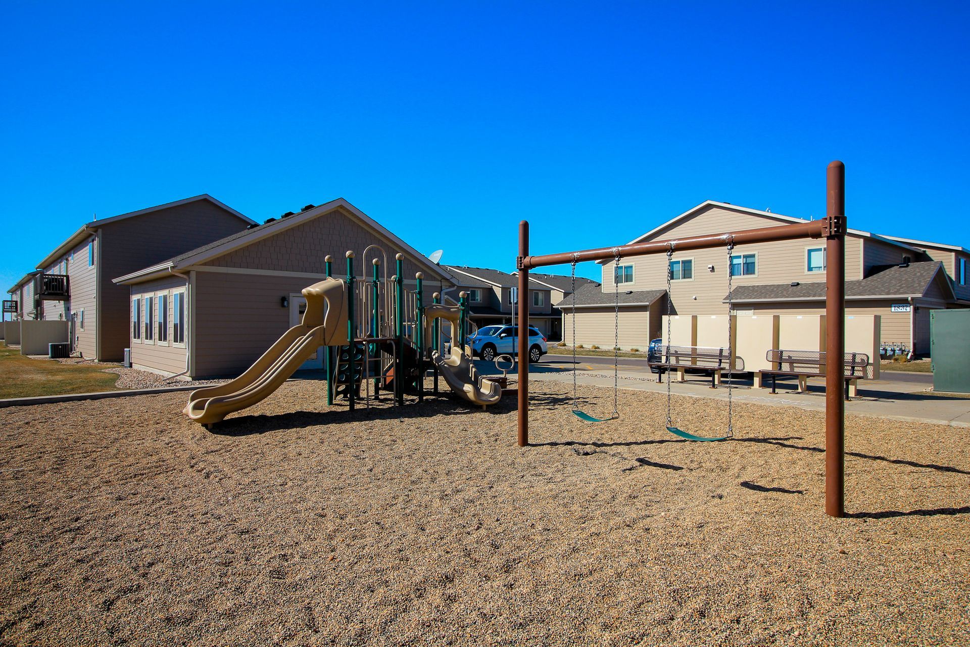Playground with slides and climbing bars on gravel beside apartment buildings under a clear blue sky