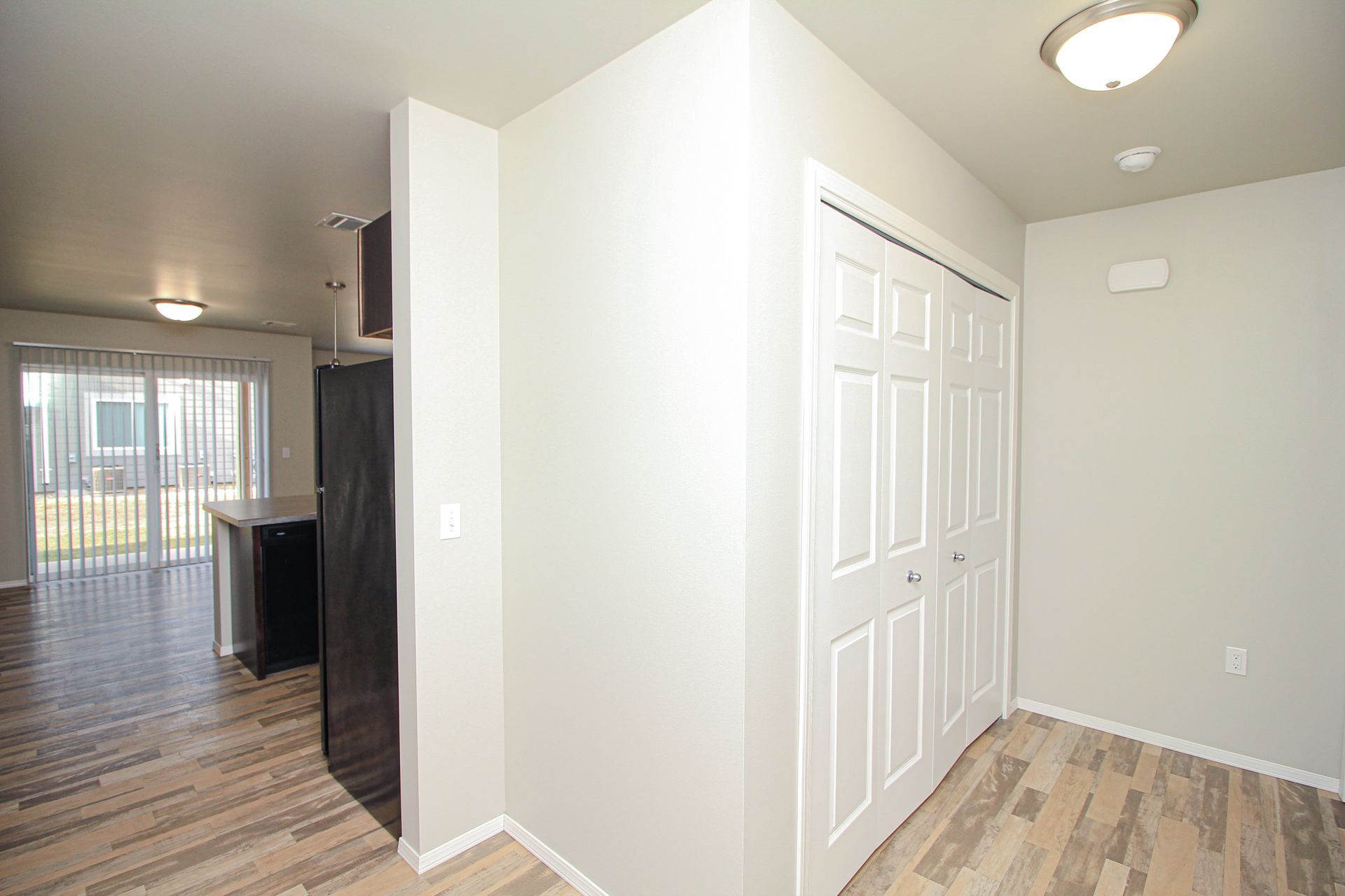 Empty hallway with light wood flooring, white closet doors, and a view into a living area and sliding glass door.