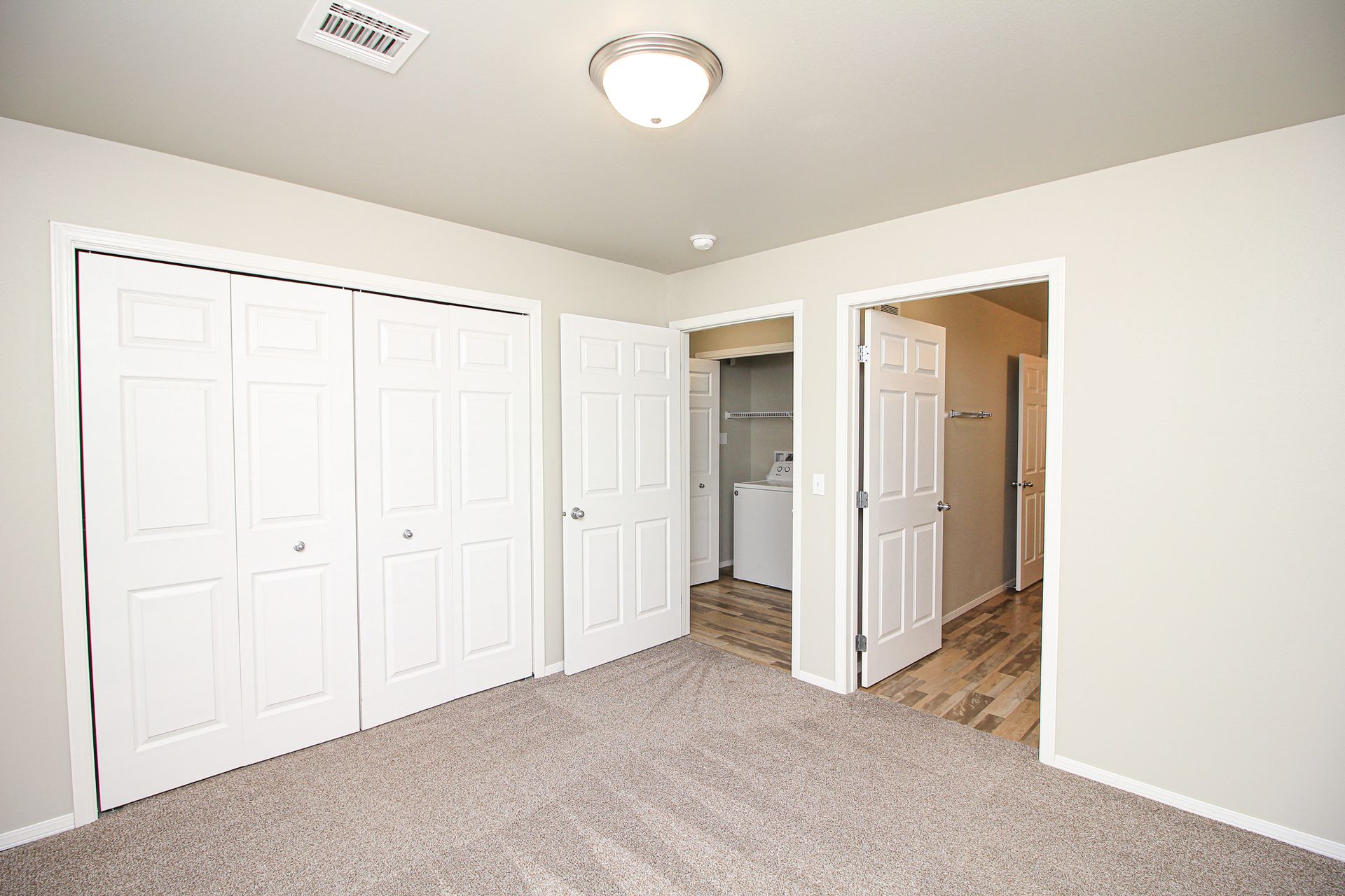 Empty beige bedroom with carpet, double closet doors, and an open doorway to a bathroom and hall