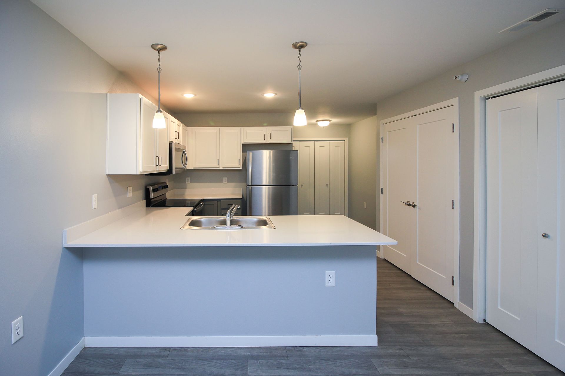 Modern white kitchen with breakfast bar, stainless steel fridge, pendant lights, and gray flooring