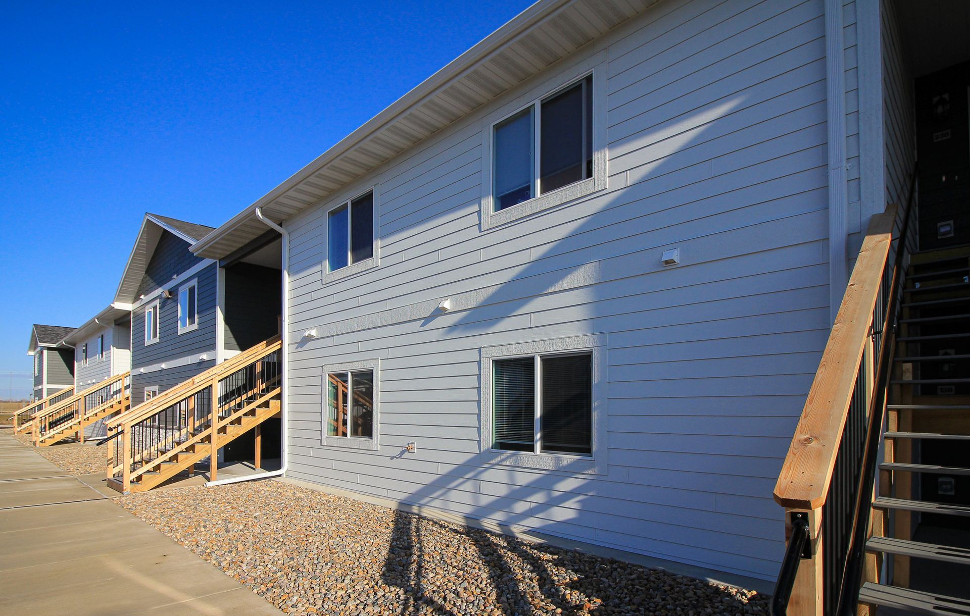 Row of white apartment buildings on a sunny slope with wooden stairways and gravel landscaping