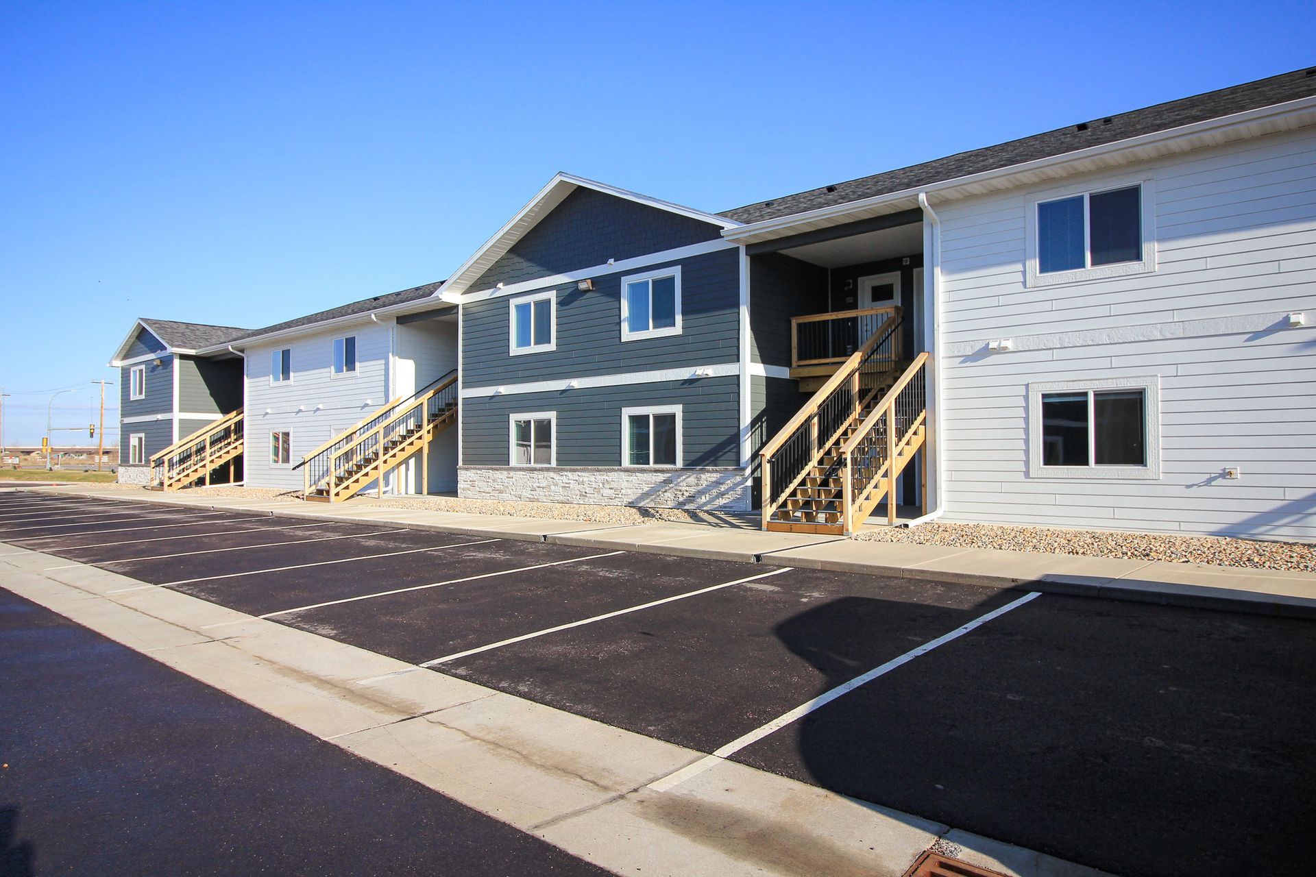 Row of newly built apartment units with white and blue siding, front porches, and parking spaces in front.