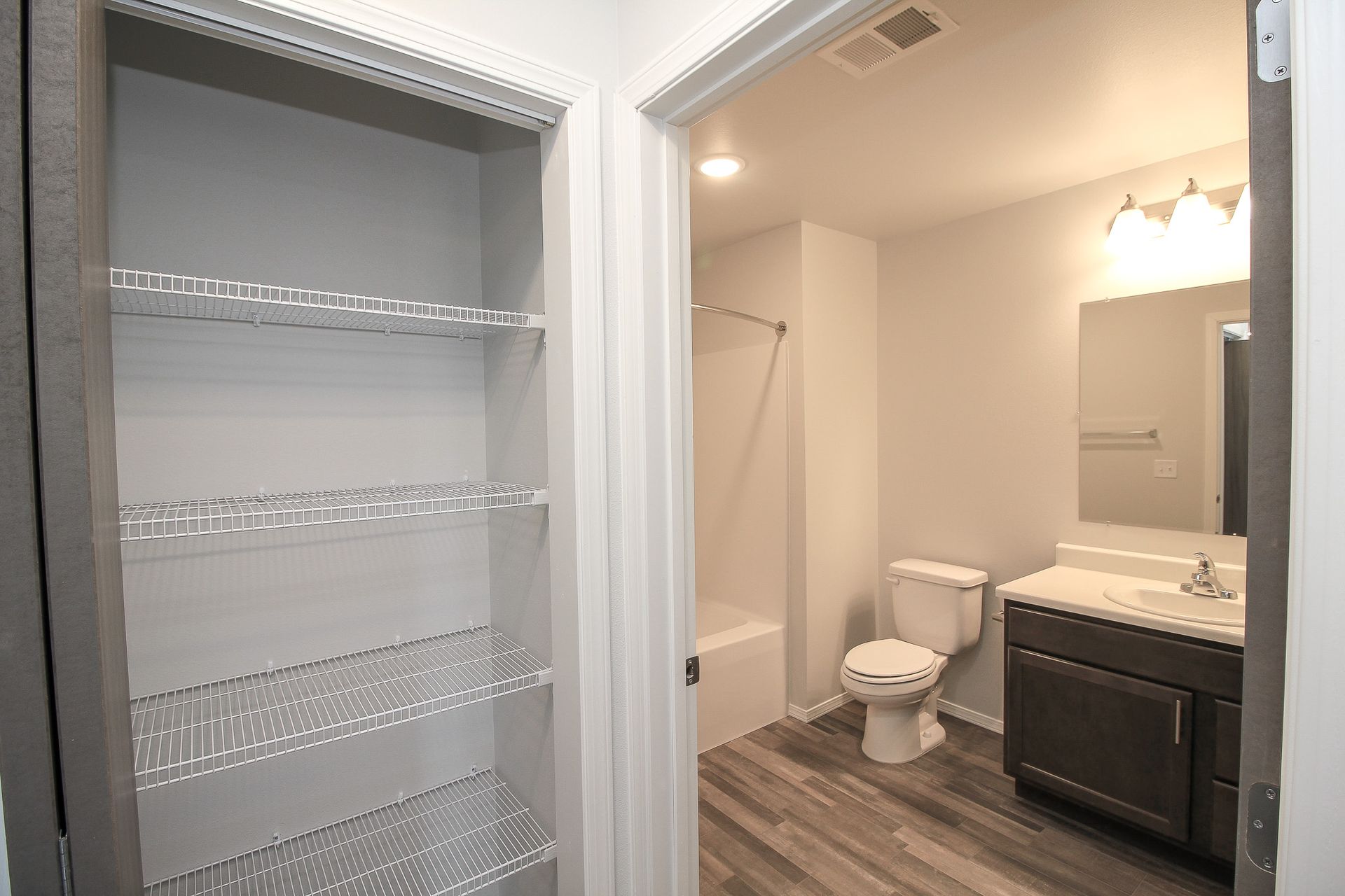 Open bathroom with shower, toilet, dark vanity, and mirrored sink beside a closet shelf.