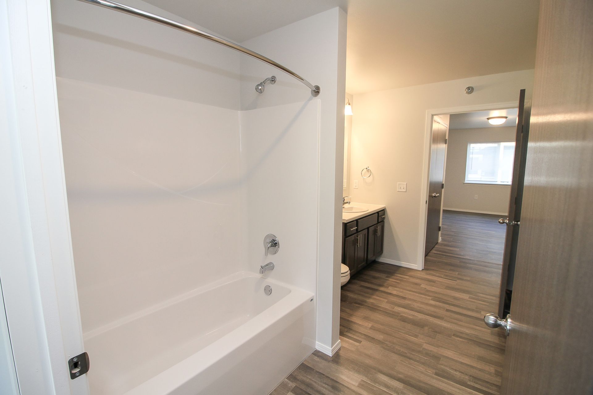 Empty white shower stall beside a hallway with wood-look flooring and a bright room beyond