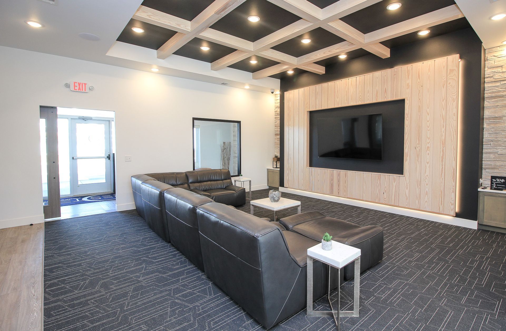 Modern lounge with black sofas, patterned carpet, and a large wall-mounted TV on a wood accent wall