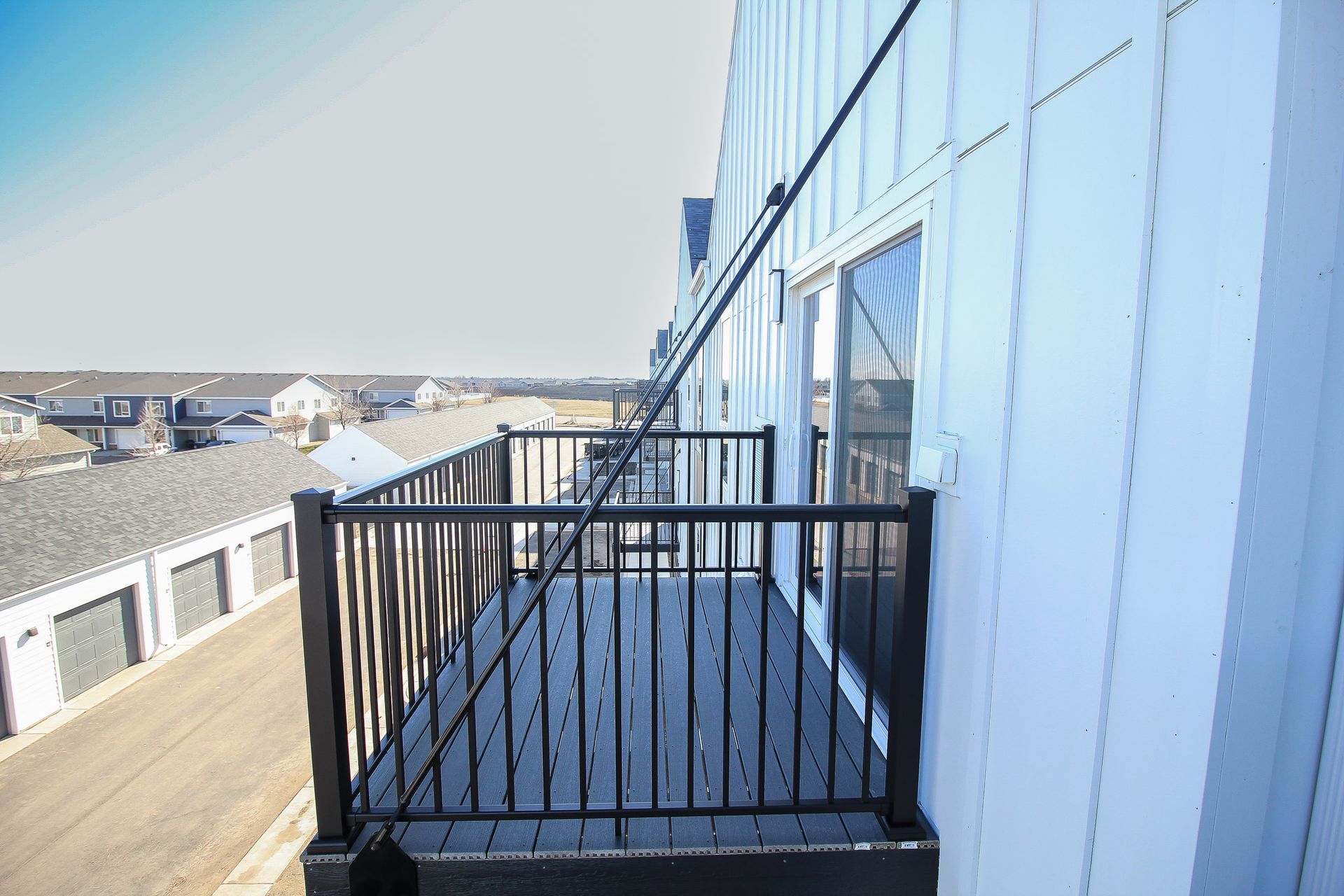 Narrow outdoor balcony with black railing beside a light blue building, overlooking rooftops and the street