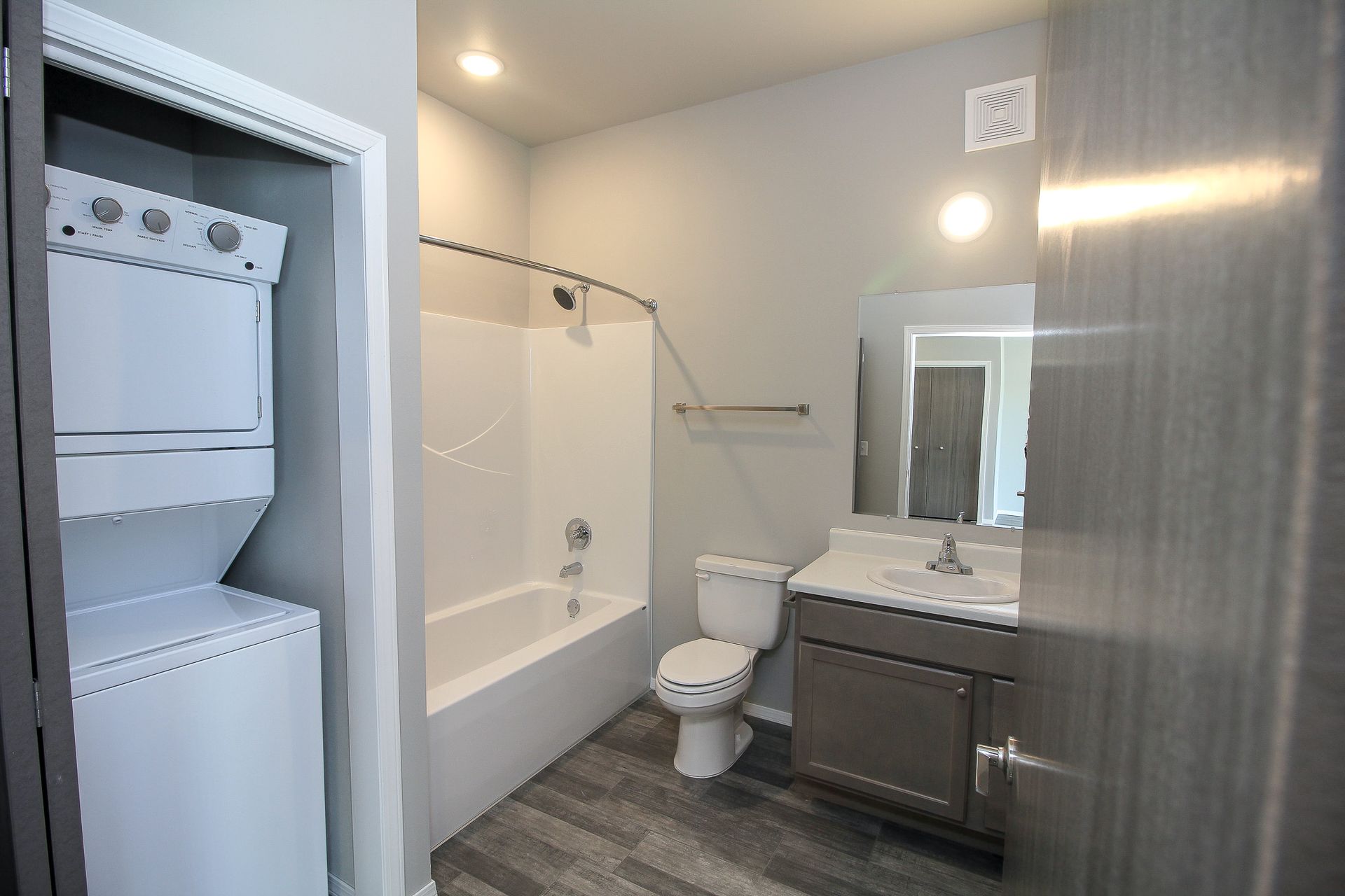 Compact bathroom with stacked washer-dryer, bathtub, toilet, vanity, and gray wood-look floor.