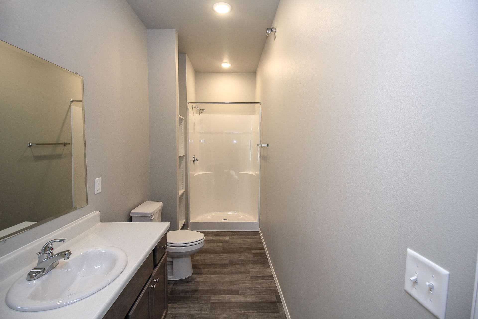 Narrow modern bathroom with sink, toilet, and shower, featuring gray walls and dark wood flooring