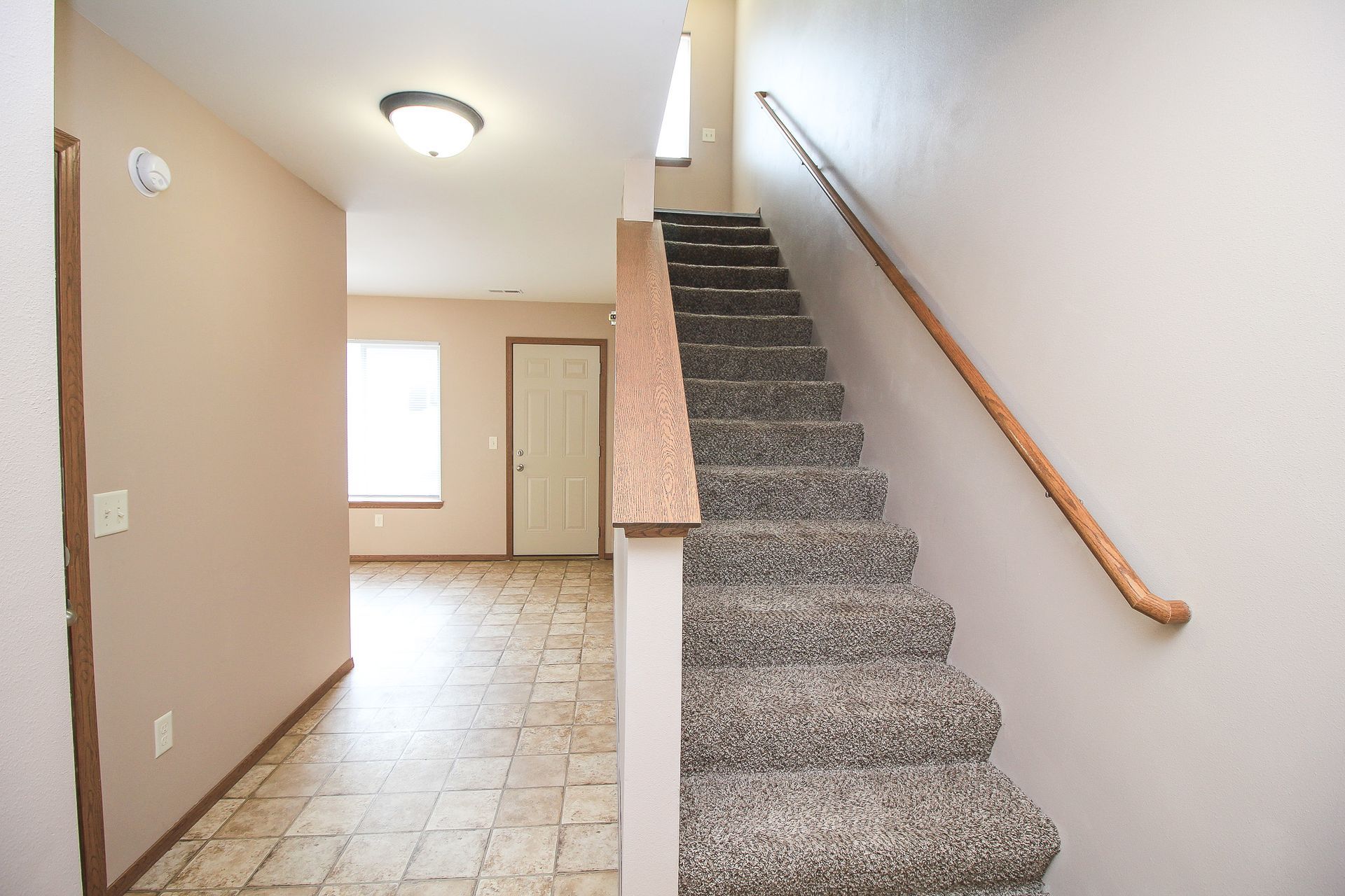 Carpeted staircase with wooden handrail in a beige entry hallway with tiled floor and front door visible