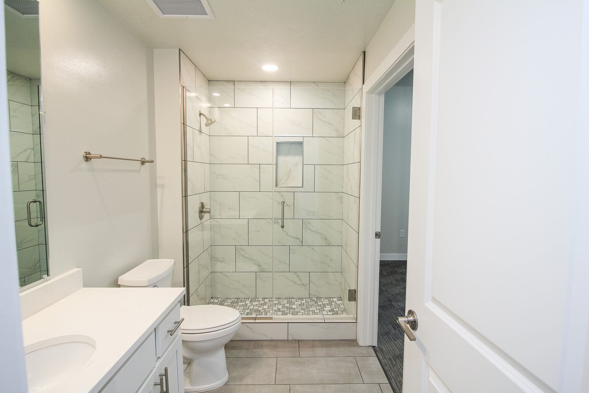 Bright bathroom with a toilet, vanity, and glass-enclosed shower with tiled walls and floor.