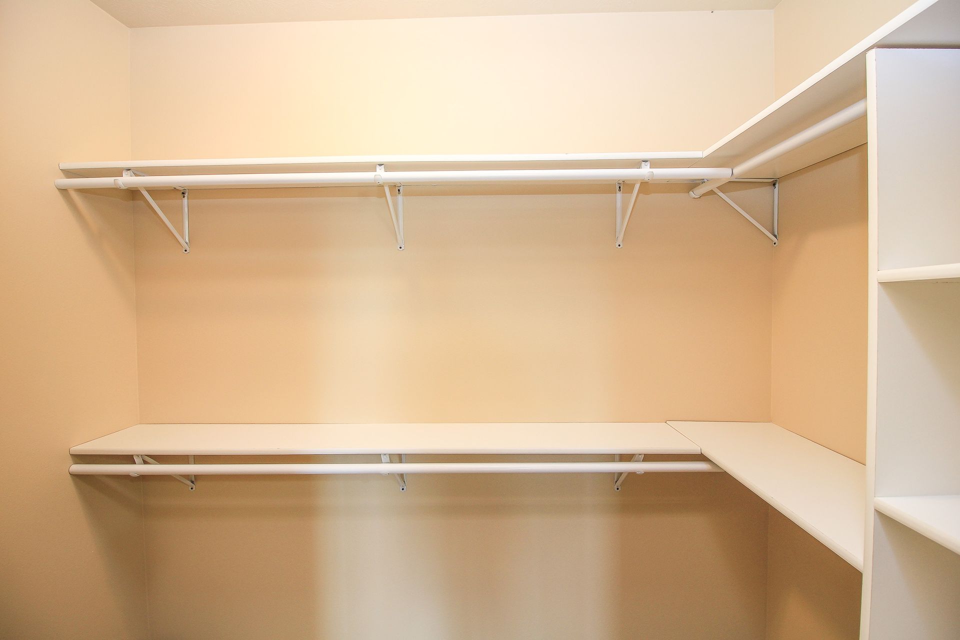 Empty walk-in closet with beige walls, white wire shelving, and hanging rods.