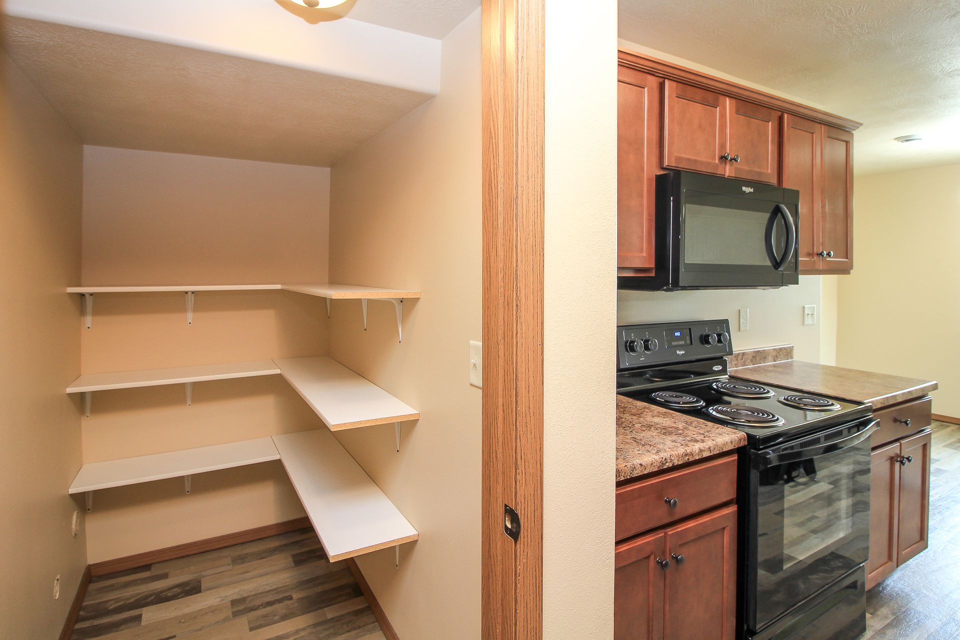 Small pantry with white shelves beside a kitchen with wood cabinets and a black stove.