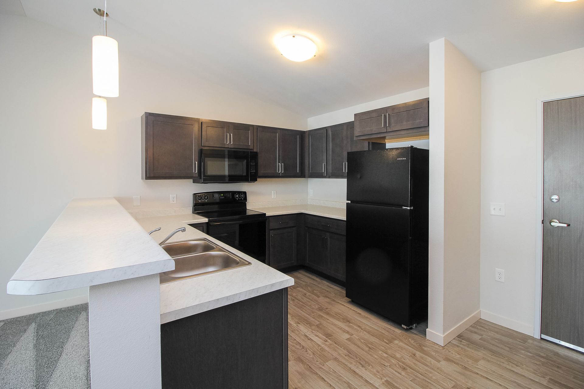 Modern kitchen with dark cabinets, black appliances, white countertops, and wood-look flooring.