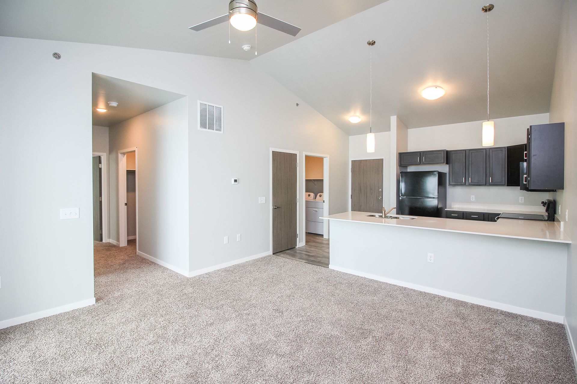 Bright open living room with carpet, white walls, ceiling fan, and a kitchen with dark cabinets and island.
