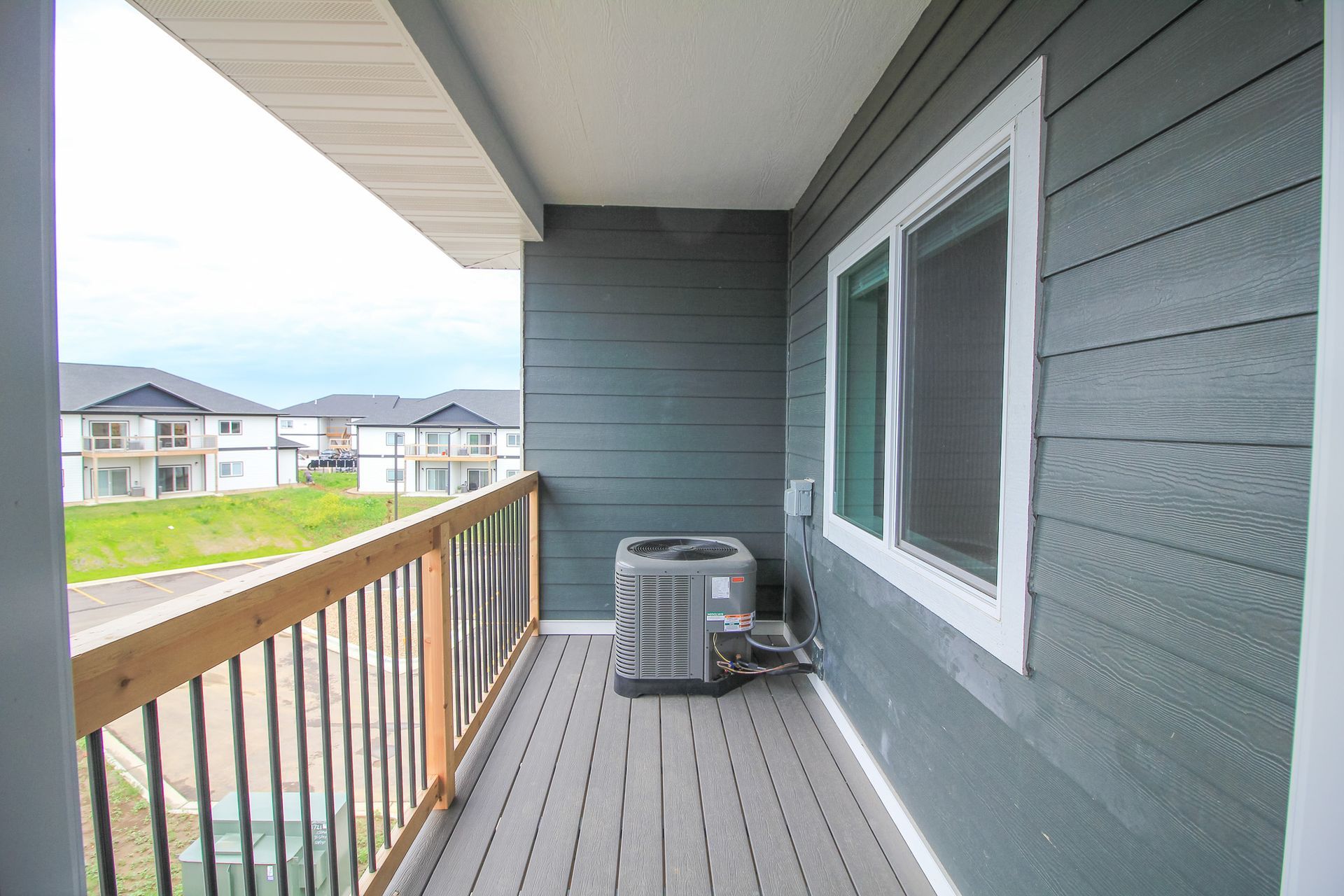 Apartment balcony with gray siding, wood railing, AC unit, and neighboring homes in view.