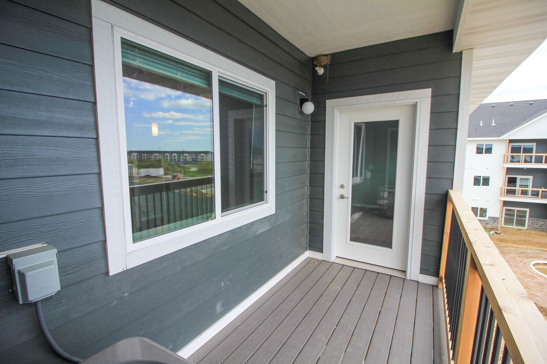 Apartment balcony with gray siding, white door and window, and wooden railing overlooking buildings and water