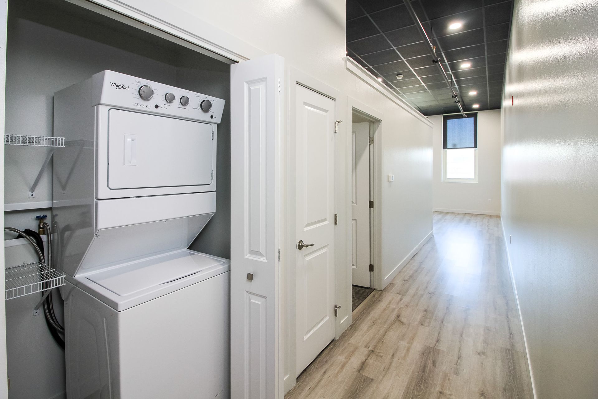 Laundry closet with stacked washer and dryer in a bright hallway with white doors and wood floors