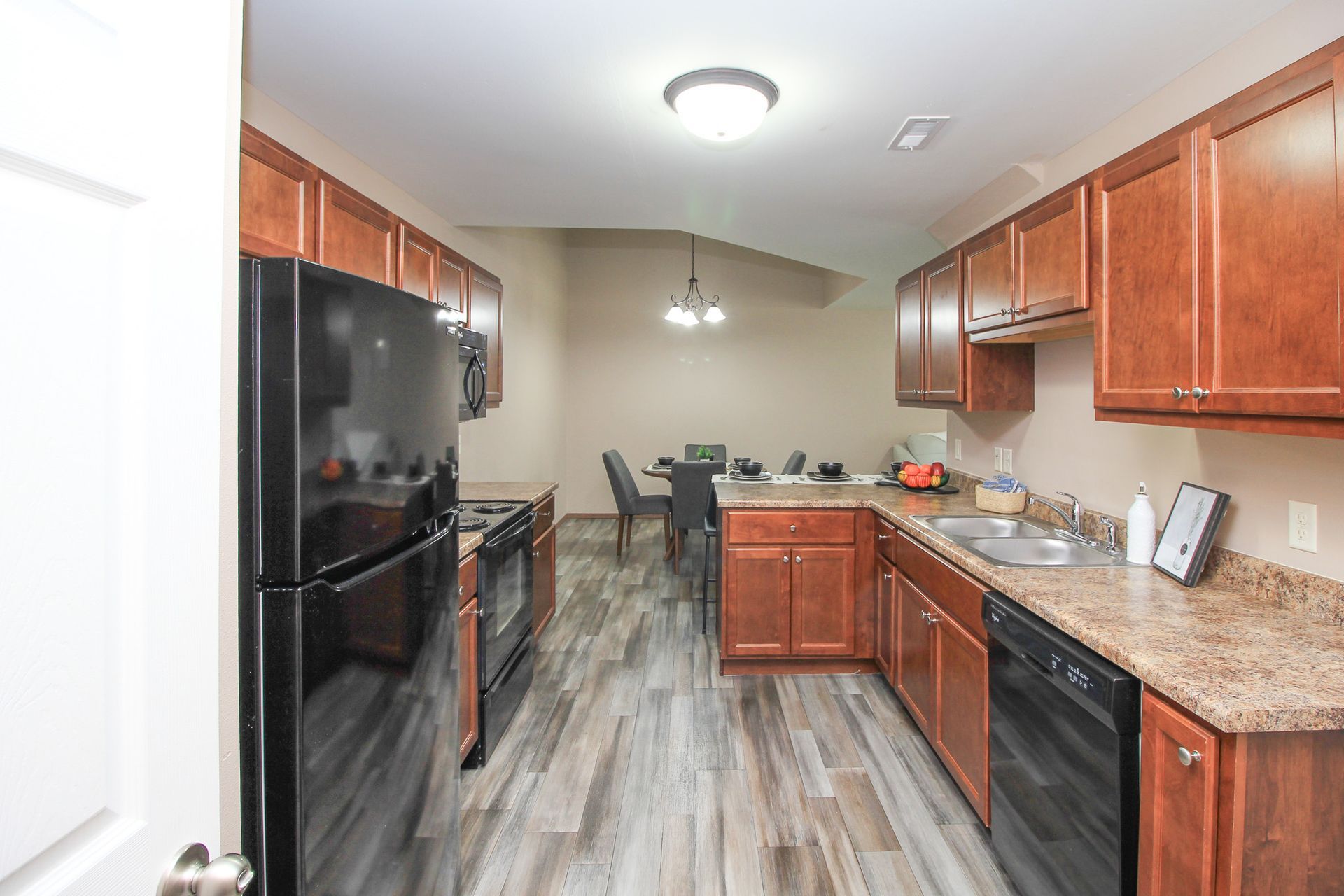 Modern kitchen with wood cabinets, black appliances, and gray wood-look flooring leading to a dining area