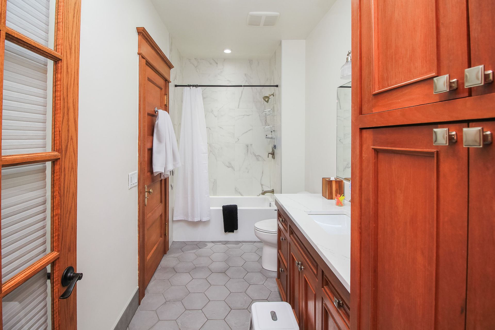 Bright bathroom with wooden cabinets, hex tile floor, white tub-shower, and a bathrobe on the door.