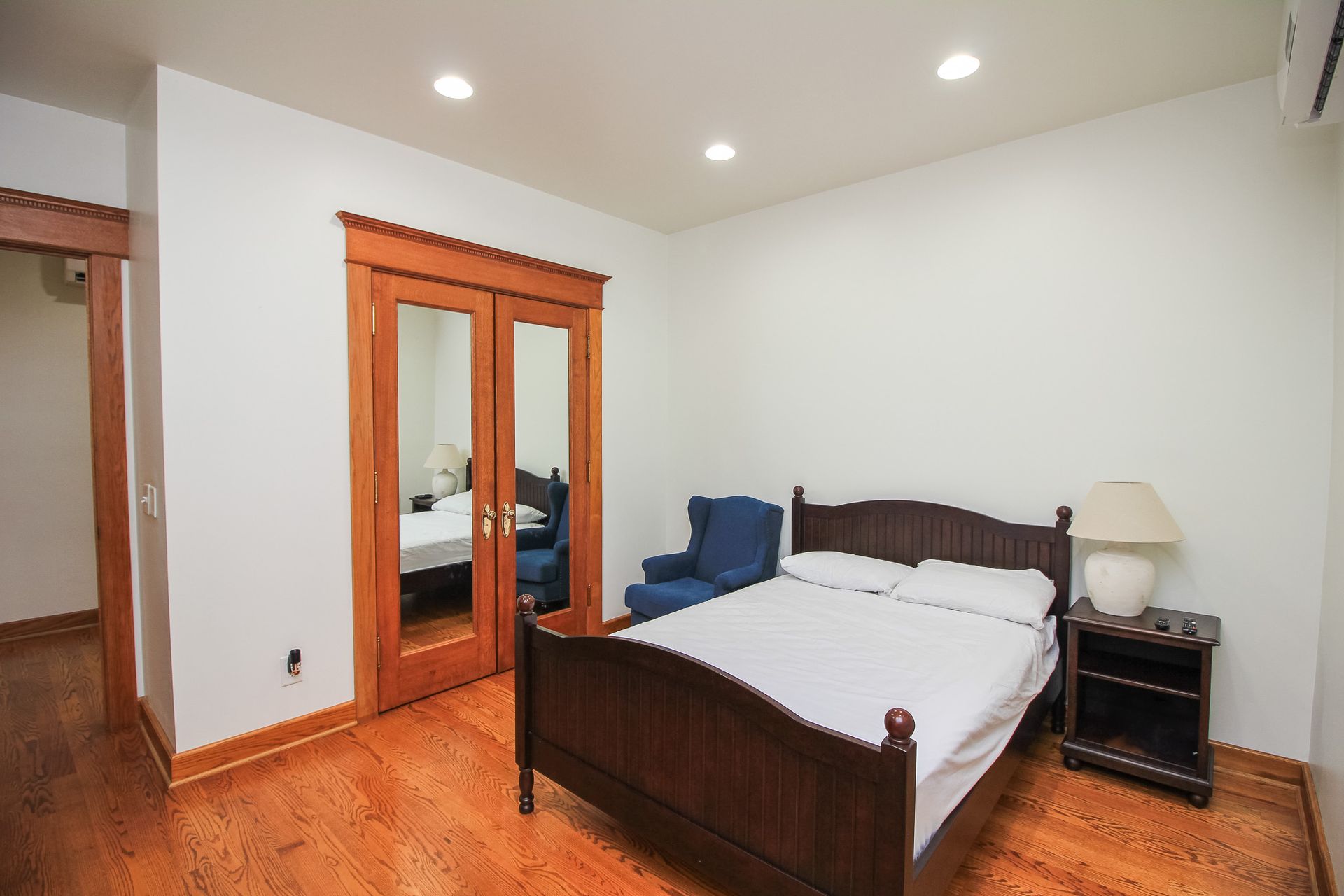 Bedroom with wooden bed, blue armchair, mirrored closet, nightstand lamp, and hardwood floors.