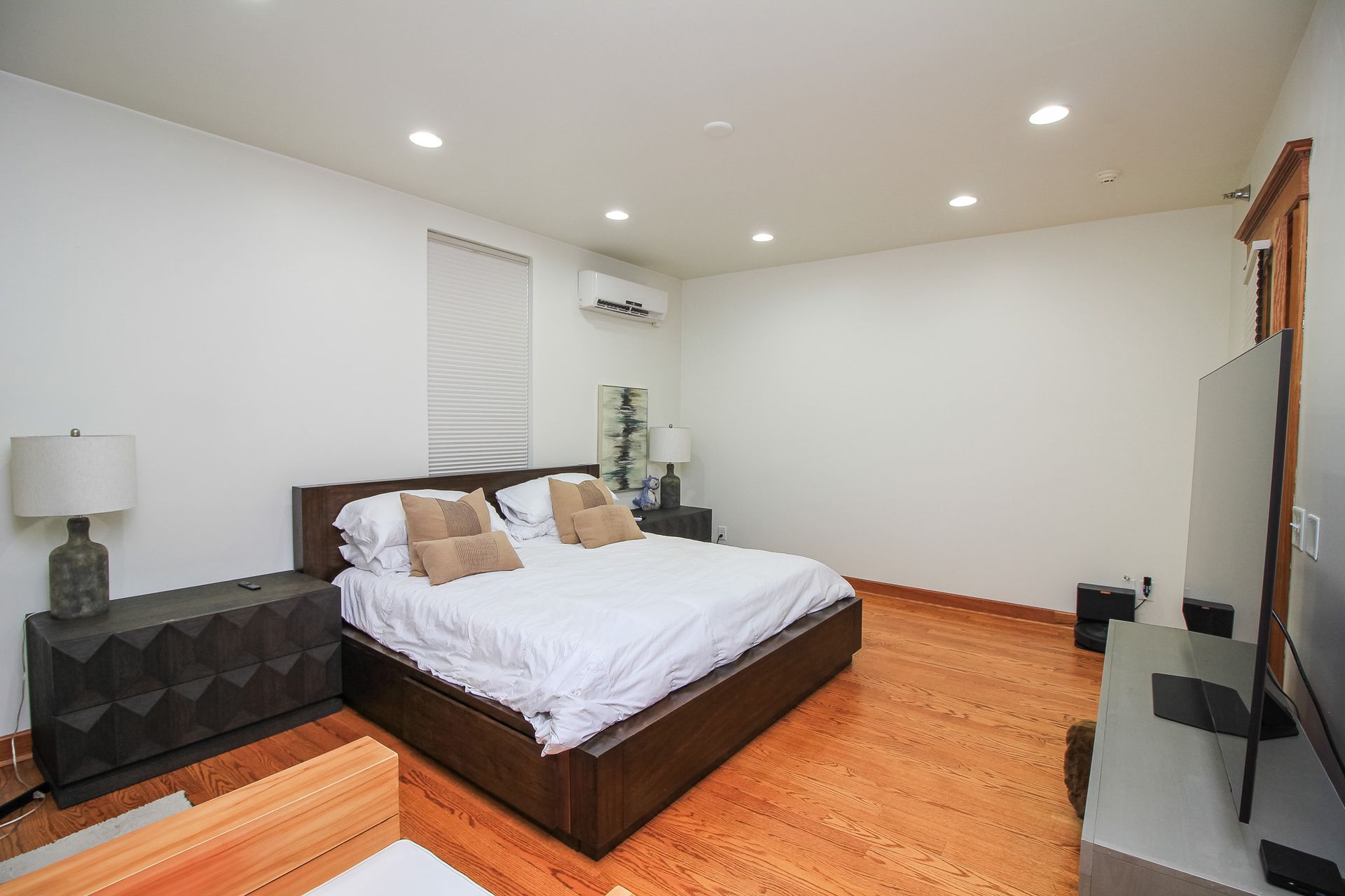 Spacious bedroom with a bed, nightstand, lamp, wall-mounted TV, and warm wood flooring.