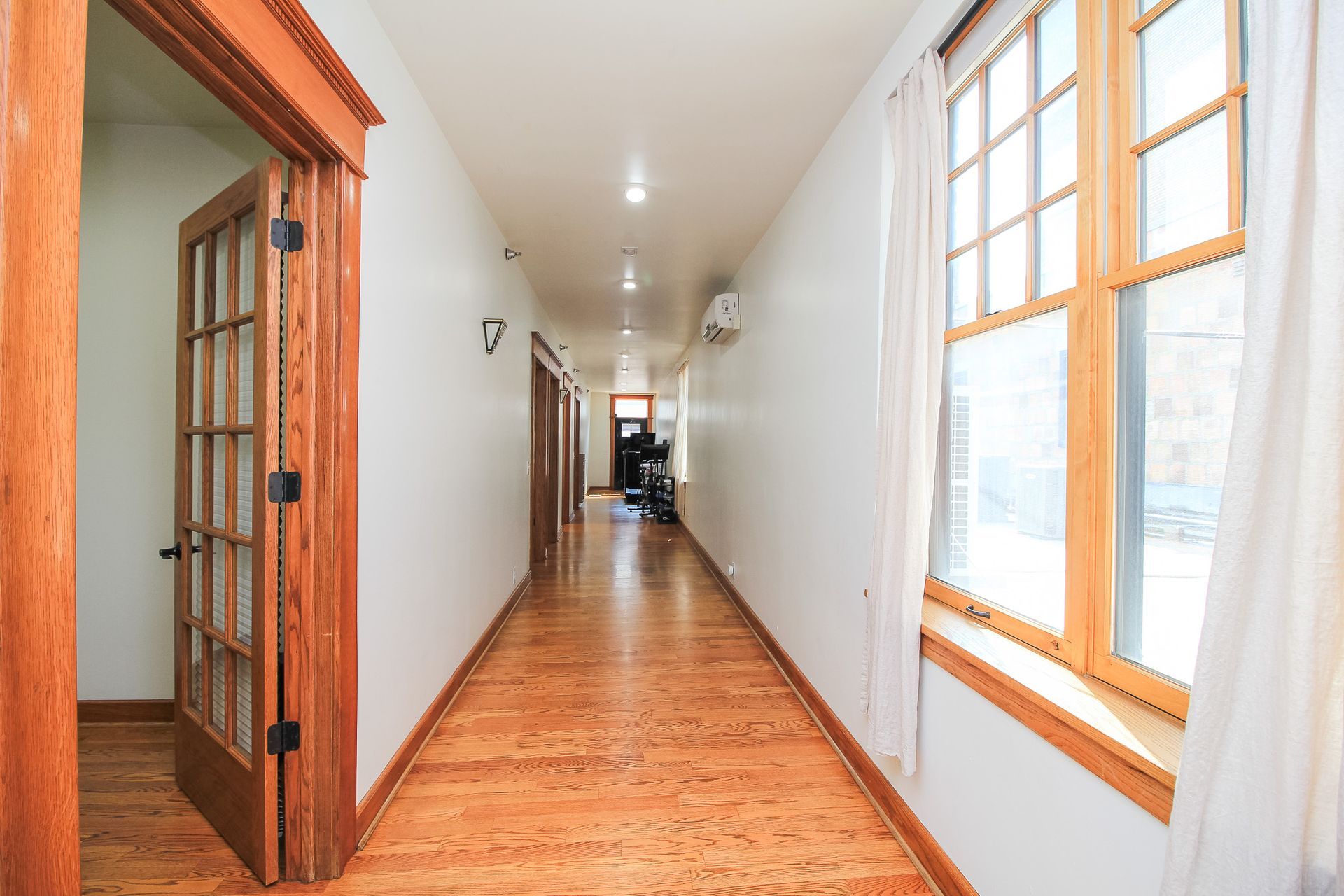 Long narrow hallway with hardwood floors, white walls, and large windows on the right