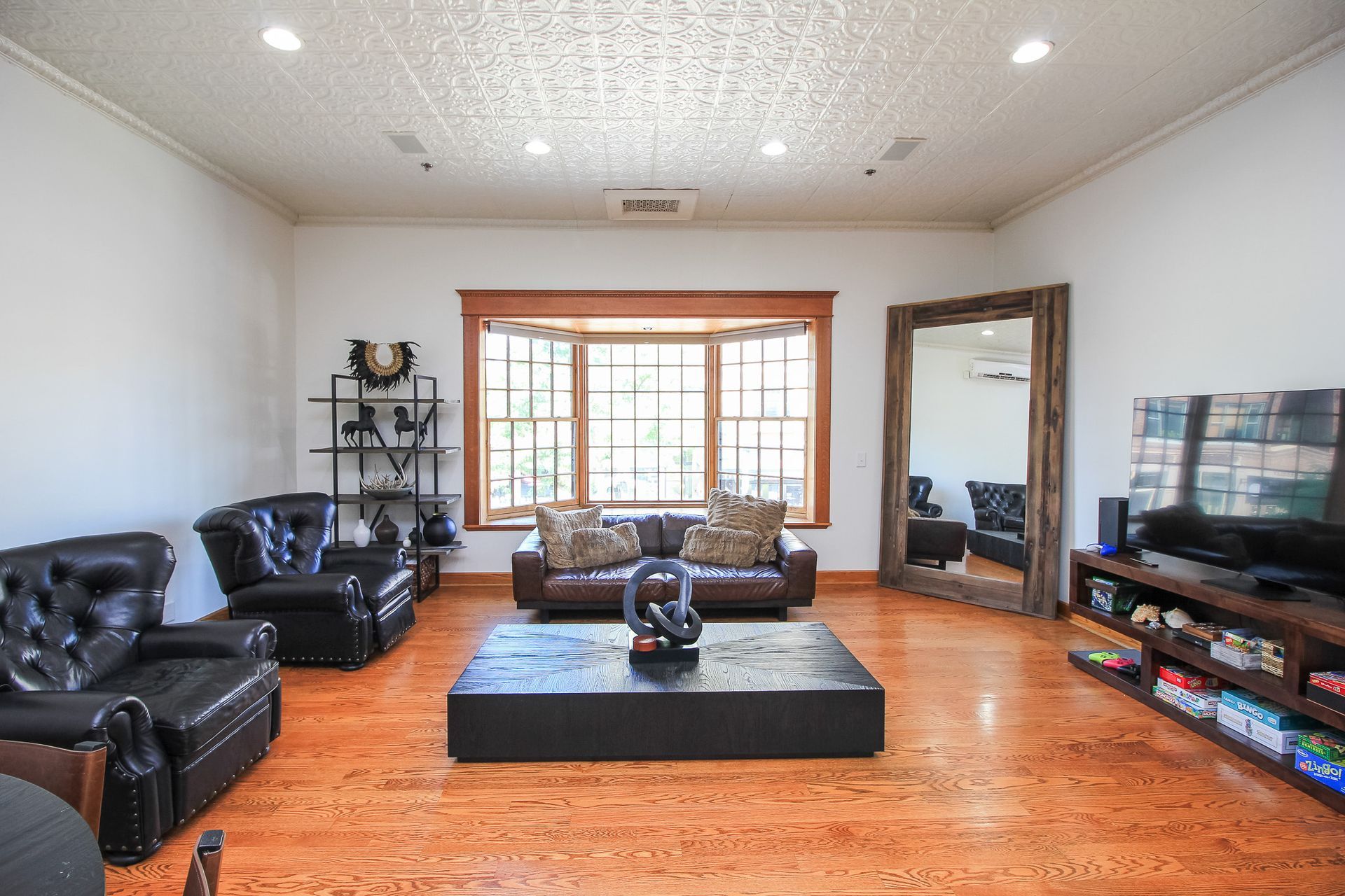 Spacious living room with black leather seating, wood floors, large window, and a TV on the right