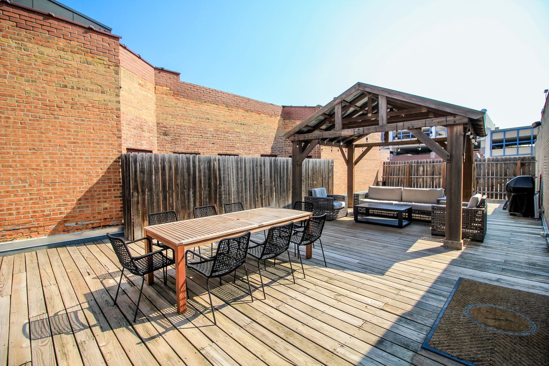 Outdoor rooftop deck with wooden tables, chairs, and a pergola beside brick walls and a city view