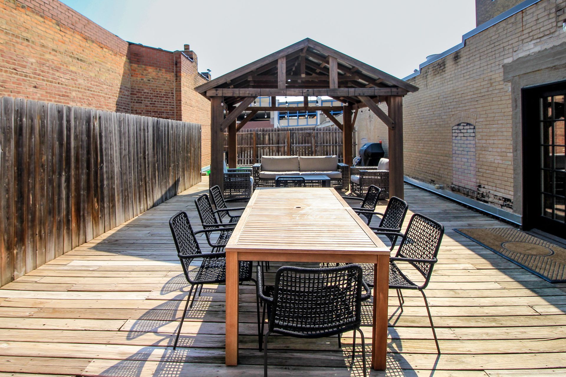 Outdoor wooden deck with long table and black chairs beside brick walls under a covered pavilion