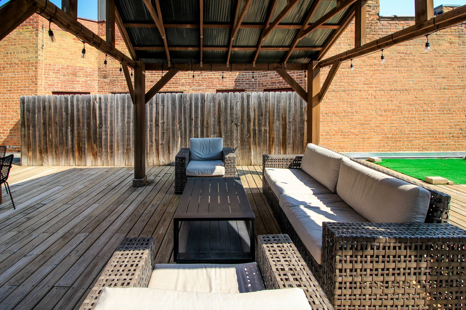 Covered patio with wicker outdoor seating, coffee table, and wooden privacy fence in bright sunlight