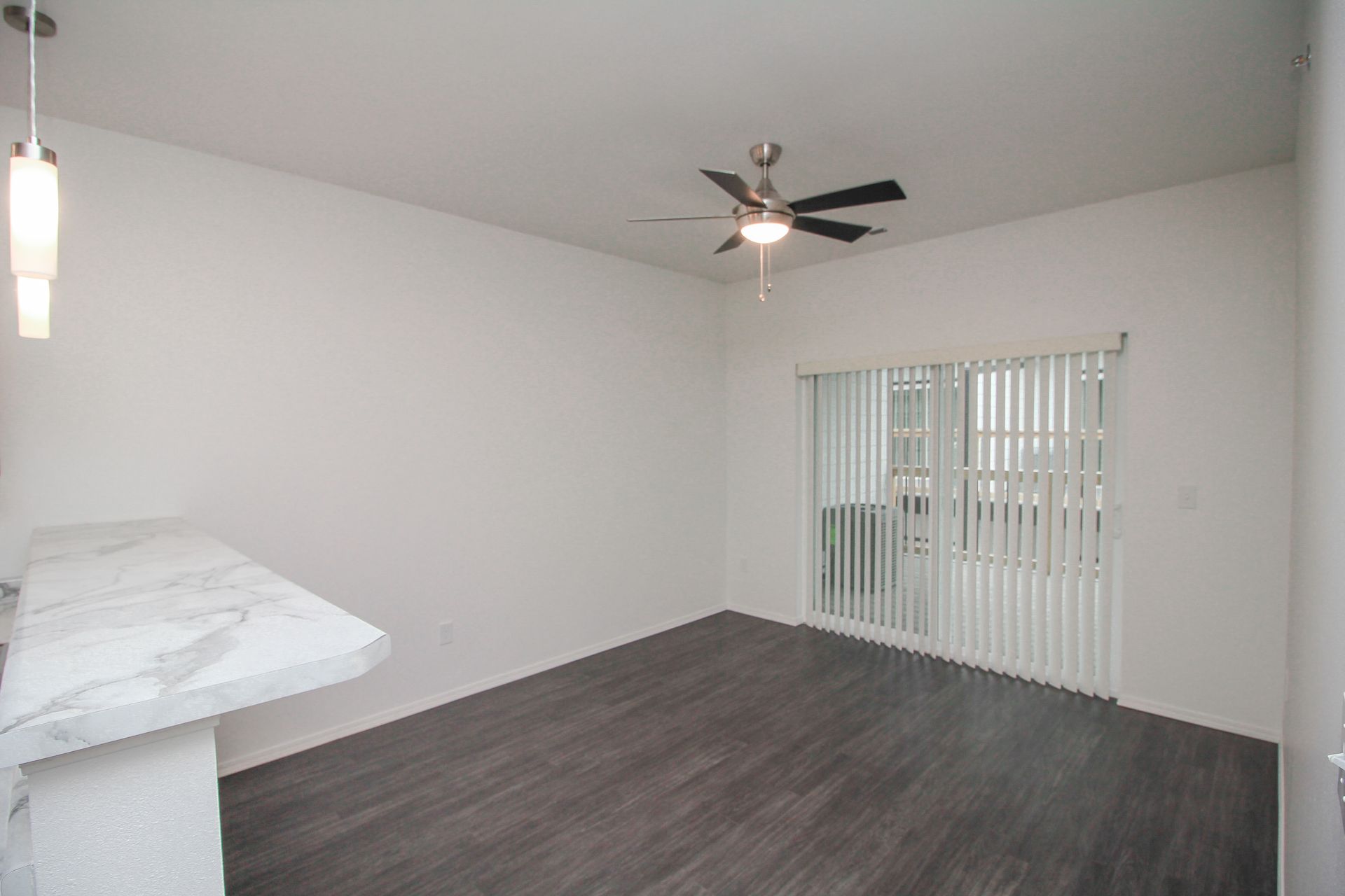 Empty modern apartment living area with white walls, gray flooring, and open kitchen with island and ceiling fan.