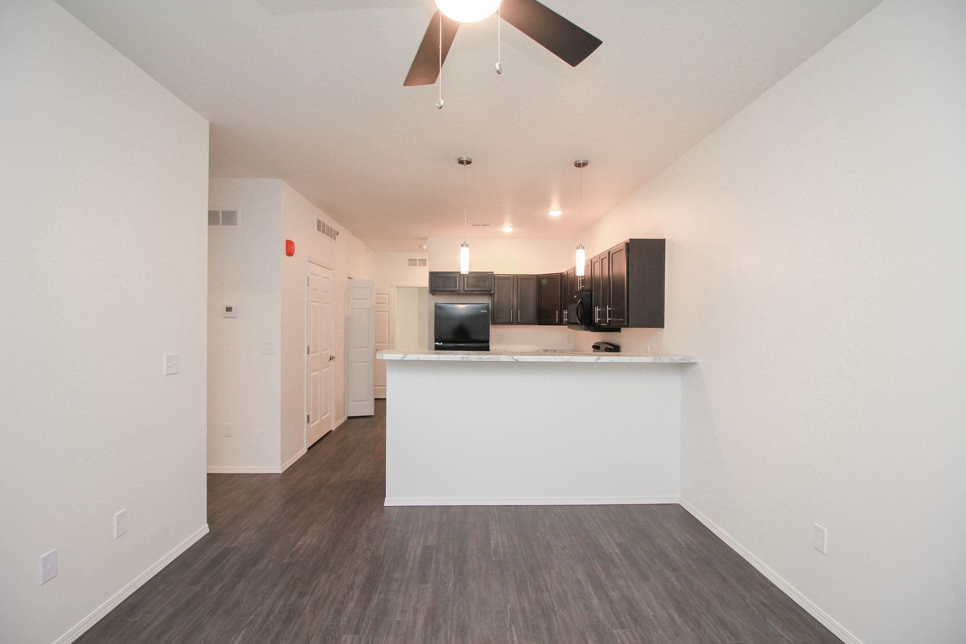 Empty white living room with gray wood floors, ceiling fan, and a window with blinds