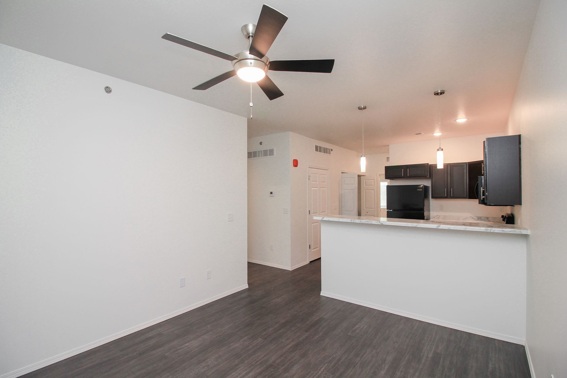 Empty apartment living room with dark flooring, white walls, black kitchen cabinets, and a ceiling fan