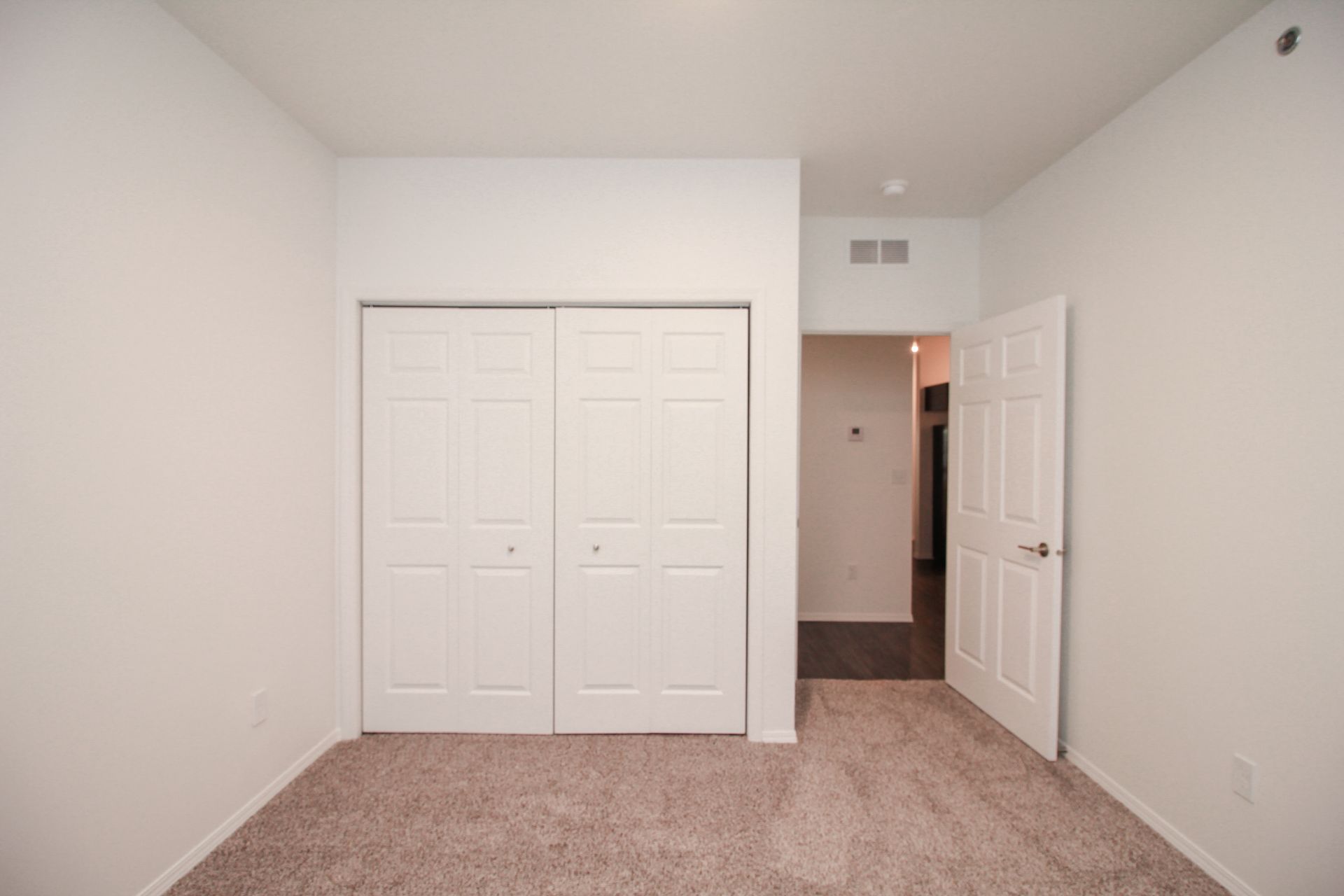 Empty bedroom with beige carpet, white walls, and double closet doors next to an open doorway.