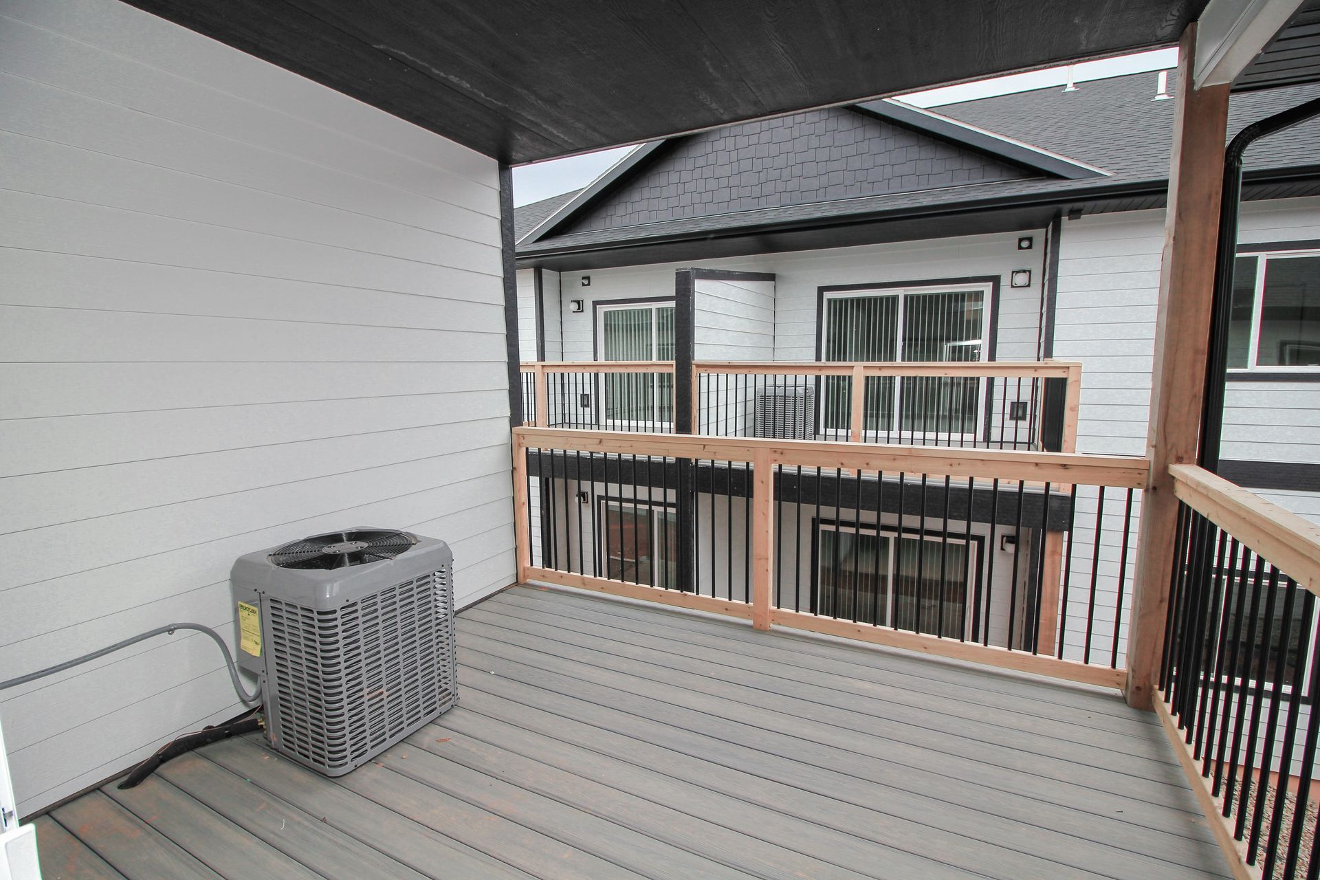 Empty covered balcony with gray flooring, white siding, wood railing, and an air conditioner unit.