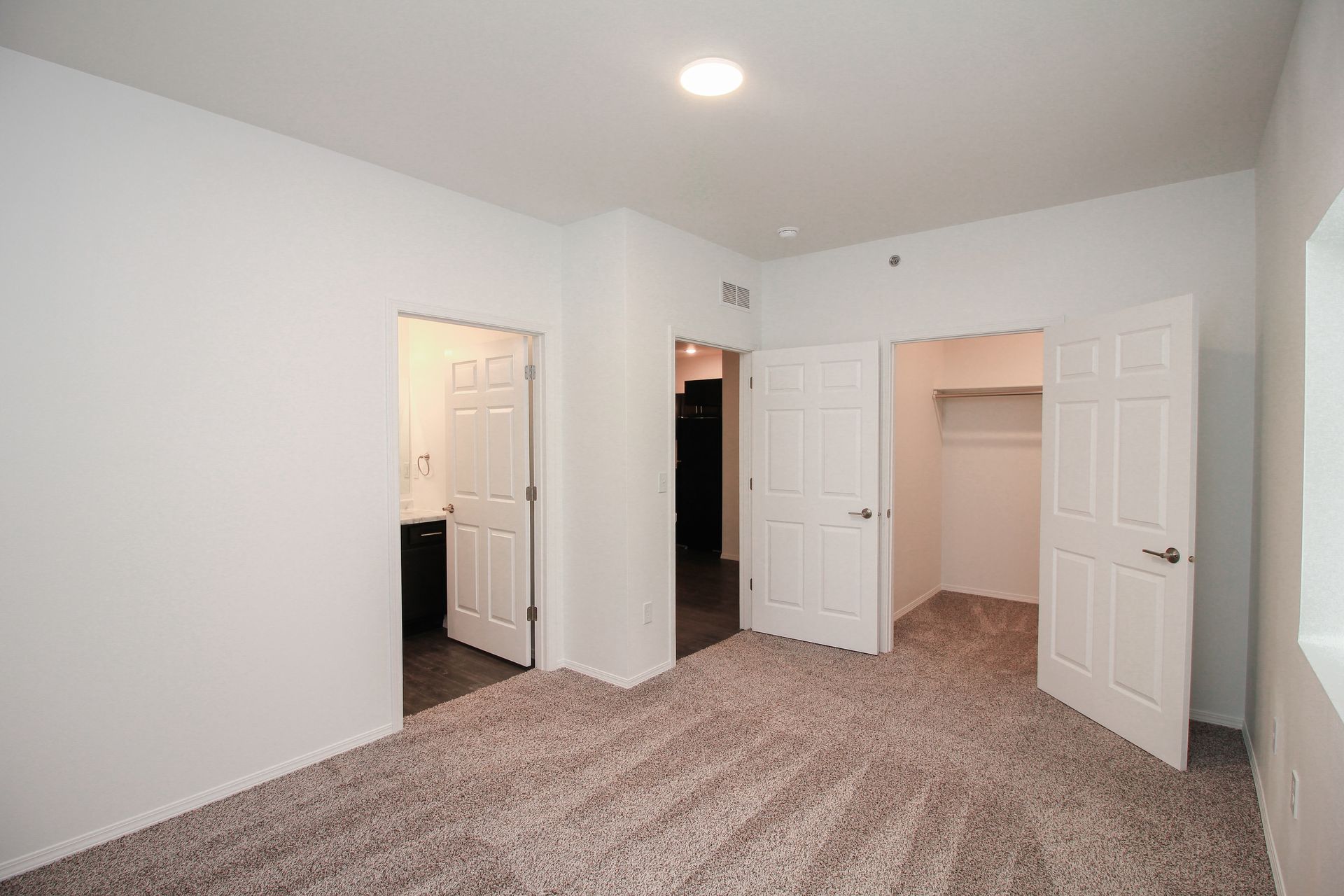 Empty carpeted bedroom with white walls, open closet doors, and a lit doorway to the hall