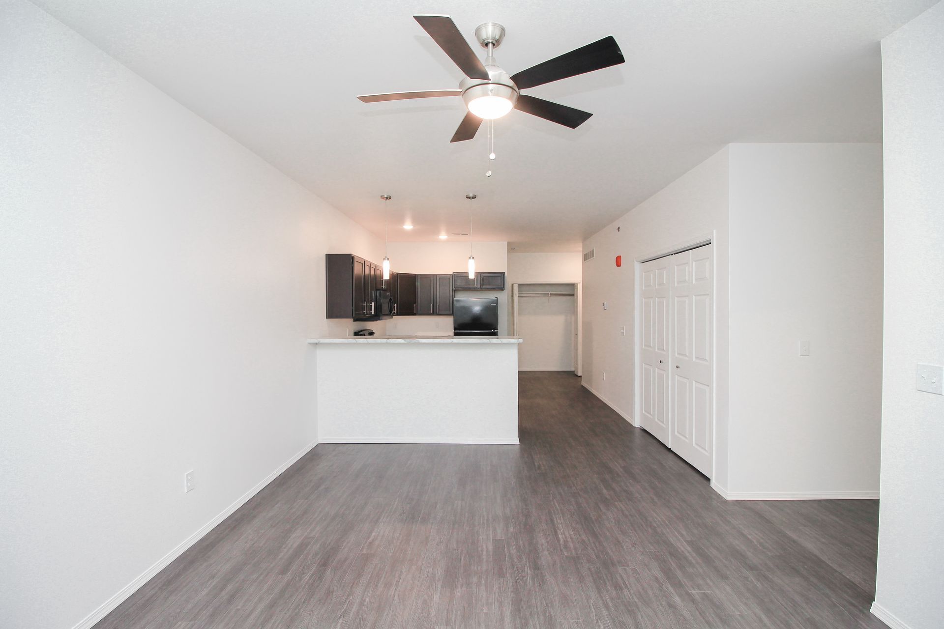 Empty modern apartment living area with gray flooring, white walls, kitchen island, ceiling fan, and closet doors