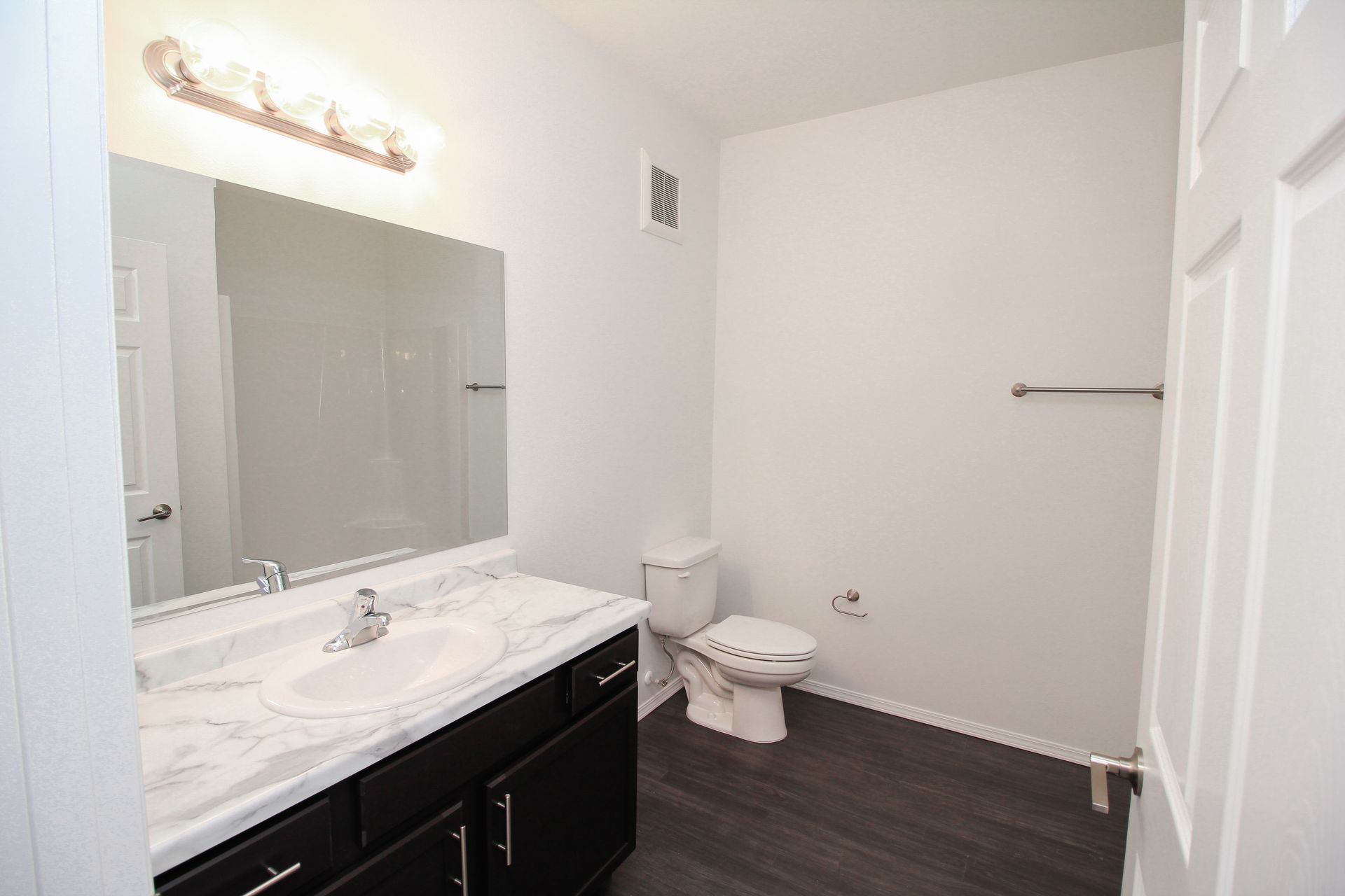 Small bathroom with a marble vanity, large mirror, white walls, toilet, and dark wood floor.